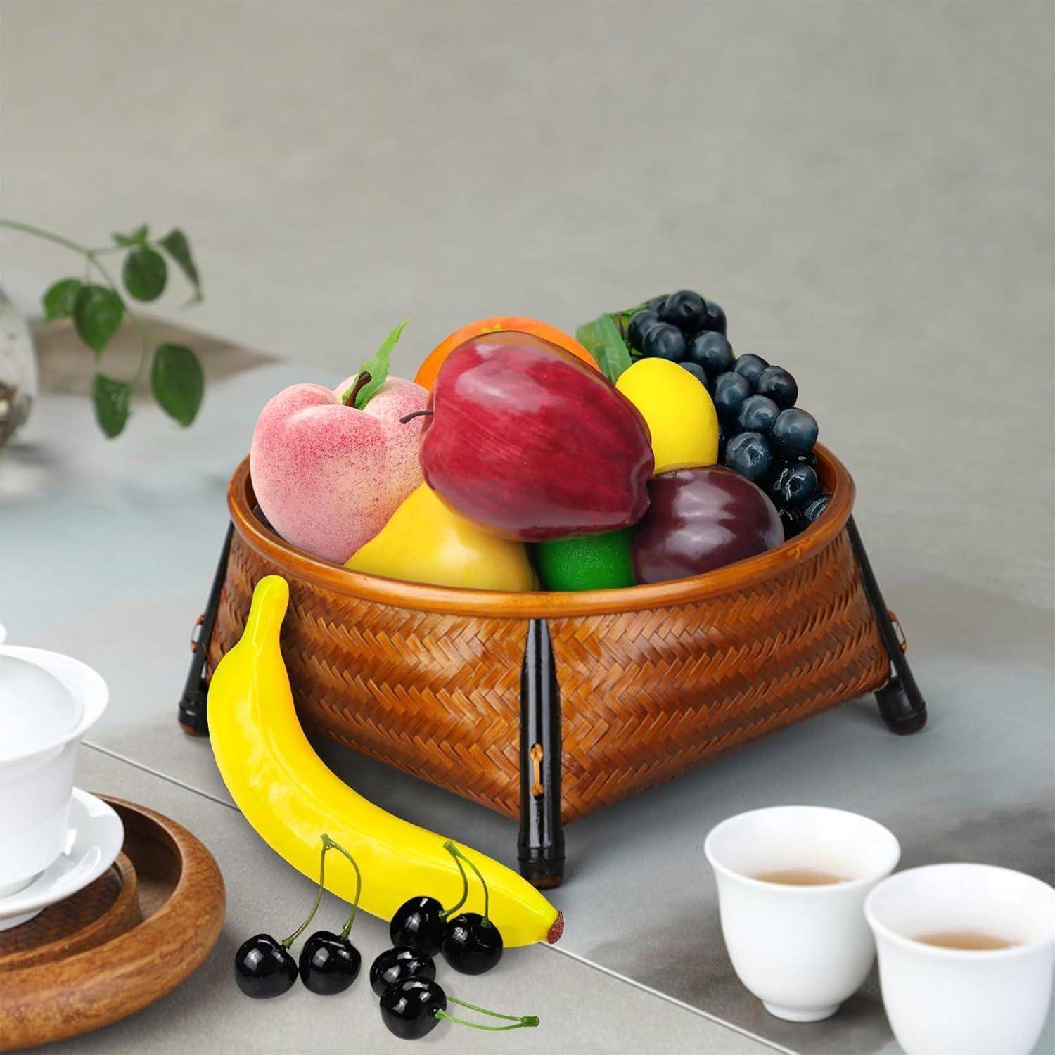 Ein geflochtener Korb enthält eine Auswahl künstlicher Früchte, darunter eine Banane, Trauben, Äpfel und eine Zitrone, die auf einem Tisch neben Teekännchen platziert sind.