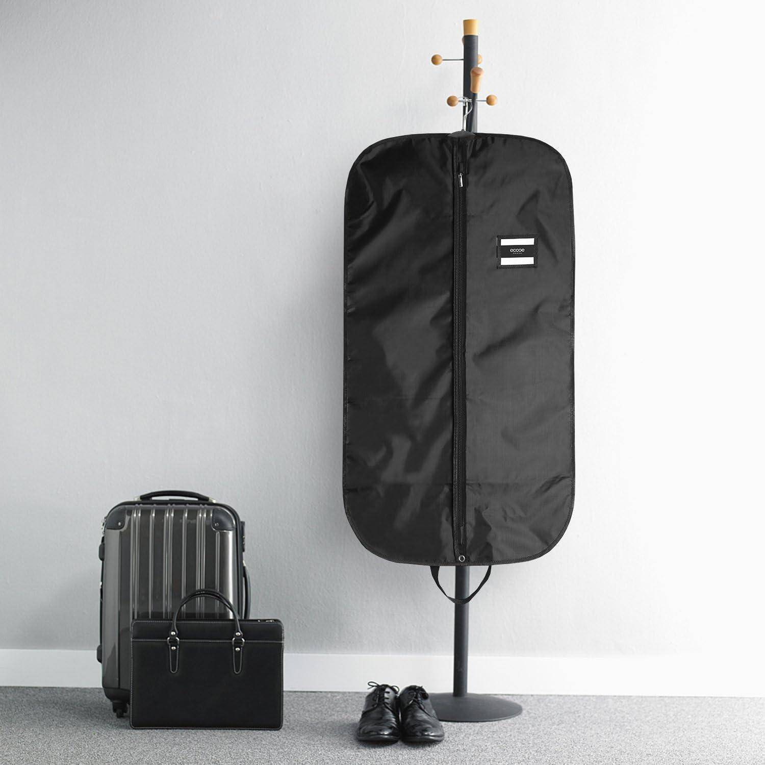 Eine schwarze Kleidertasche hängt an einem Kleiderständer neben einem silbernen Koffer, einer schwarzen Aktentasche und einem Paar schwarzer Schuhe, vor einem hellen Hintergrund.