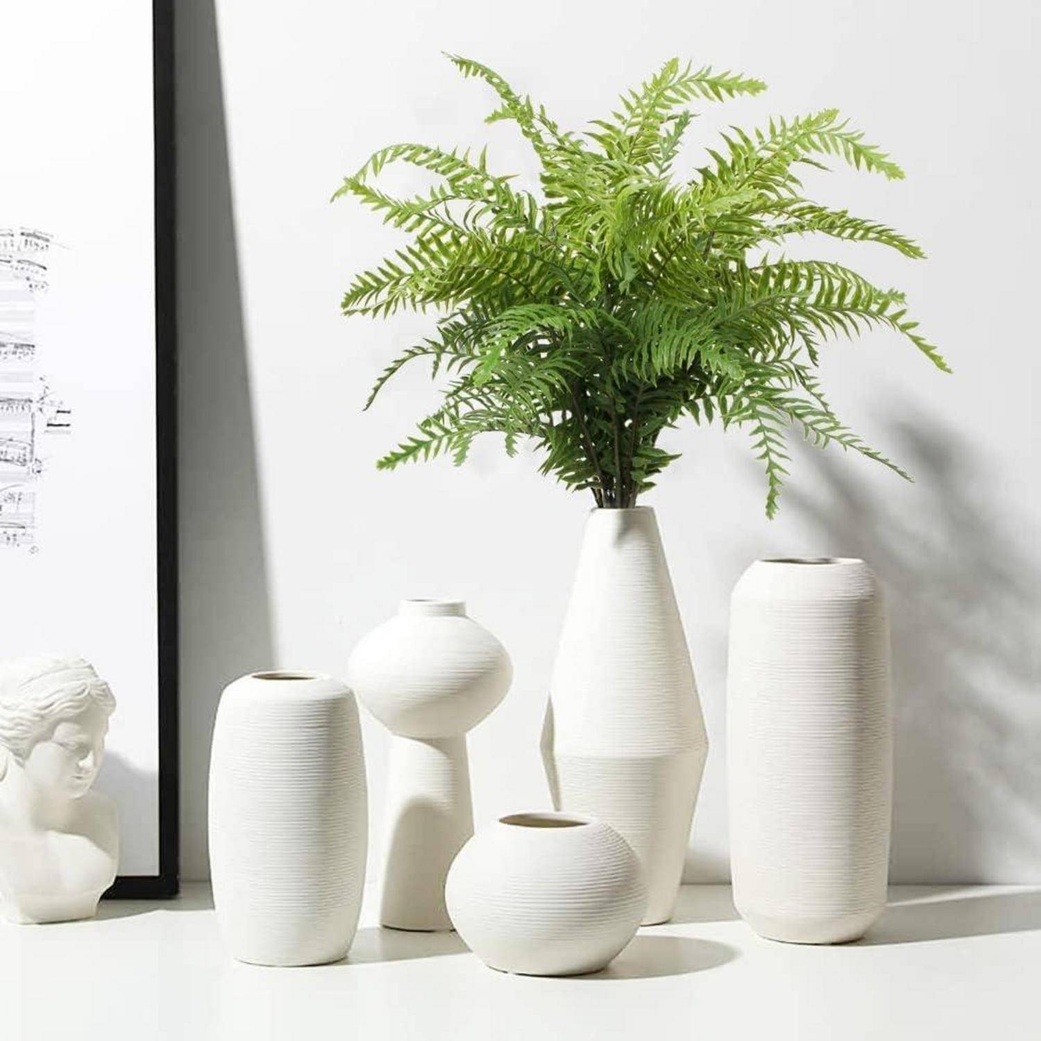 Eine Zusammenstellung aus weißen Keramikvasen und einer Büstenskulptur auf einer Oberfläche, mit einem in einer Vase platzierten Farn, vor einem teilweise sichtbaren gerahmten Kunstwerk.