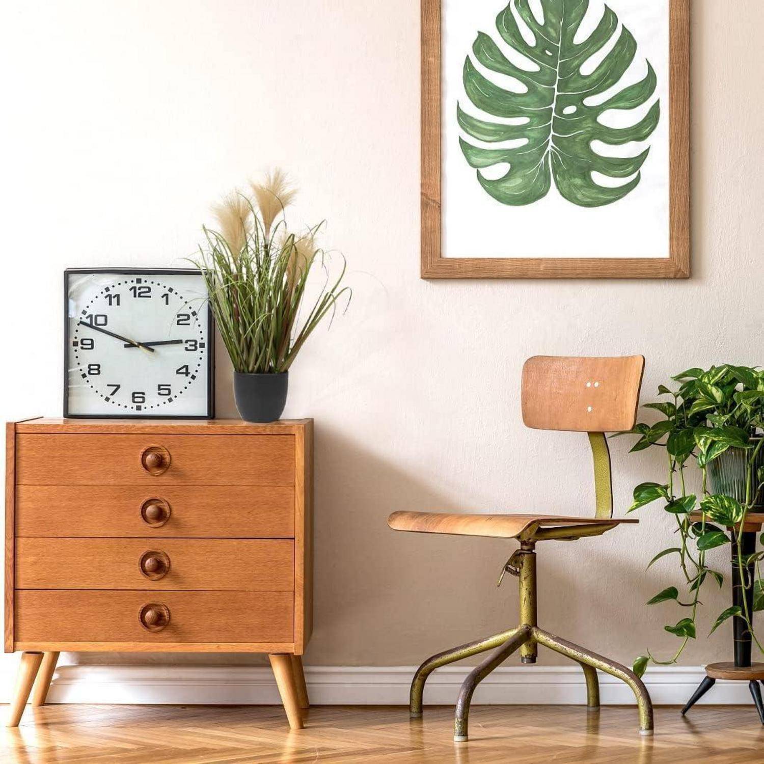 Ein gemütliches Zimmer mit einer Holzkommode, einer Uhr und Topfpflanzen neben einem Holzstuhl. Ein gerahmtes Kunstwerk mit einem Monstera-Blatt hängt an der Wand.