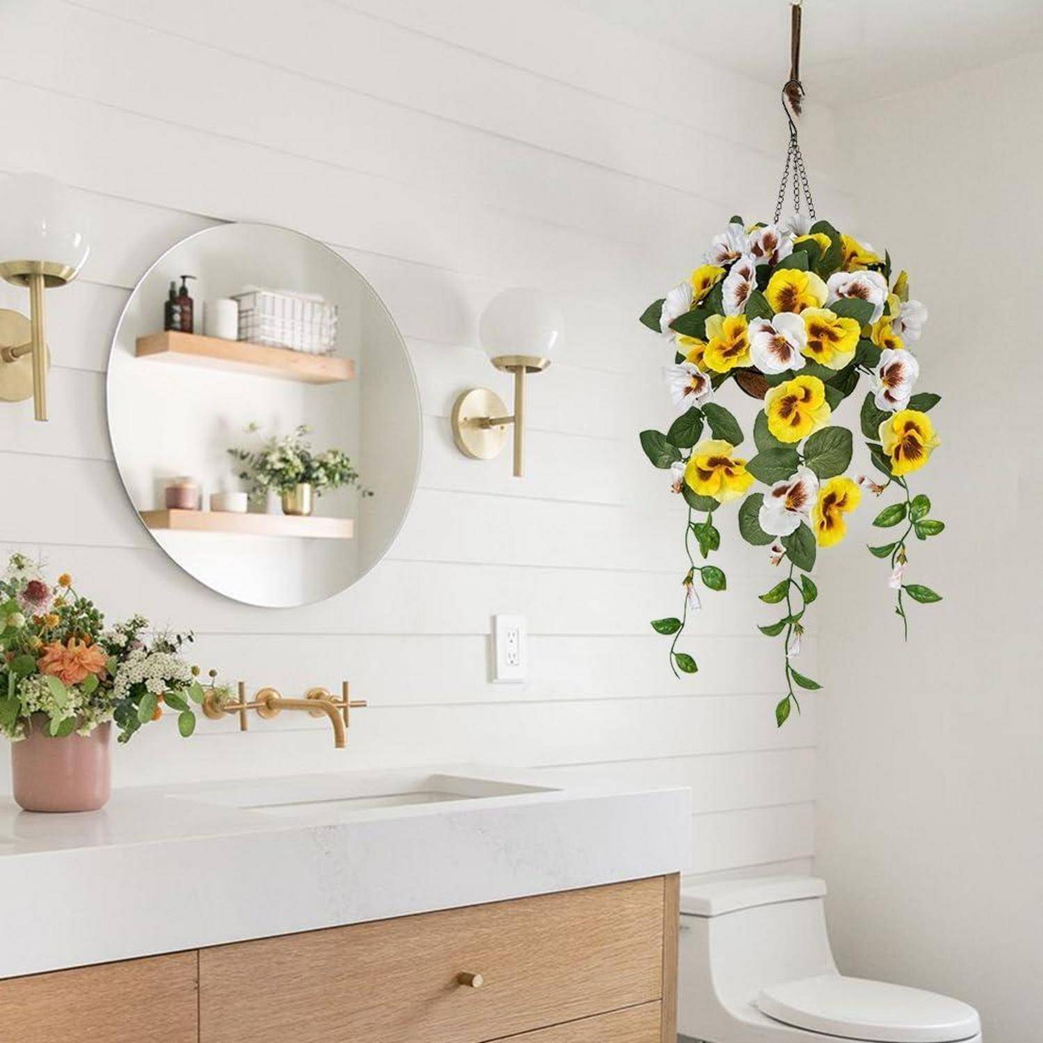 Helles Badezimmer mit weißem Waschtisch, rundem Spiegel, Topfblumen auf der Ablage und einem hängenden Korb mit lebendigen gelben und weißen Blumen.
