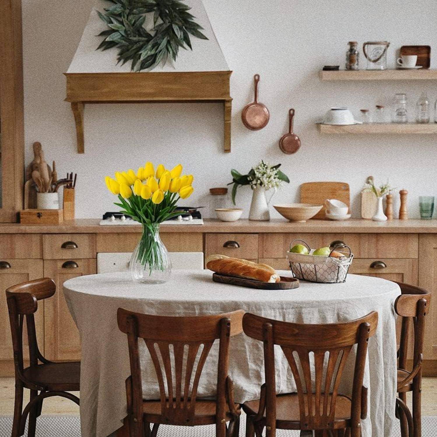 Eine gemütliche Küche mit Holzschränken, einem Tisch mit weißer Tischdecke, einer Vase mit gelben Tulpen, Brot und einer Schüssel mit Äpfeln.