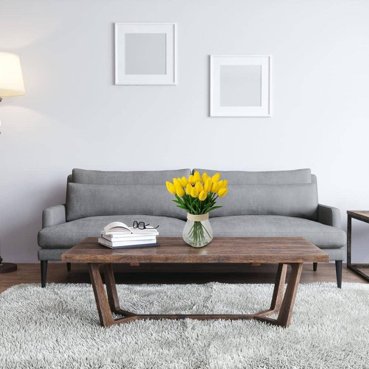 Ein modernes Wohnzimmer mit einem grauen Sofa, einem Couchtisch aus Holz mit Büchern und einer Vase mit gelben Blumen sowie zwei gerahmten Bildern an der Wand.