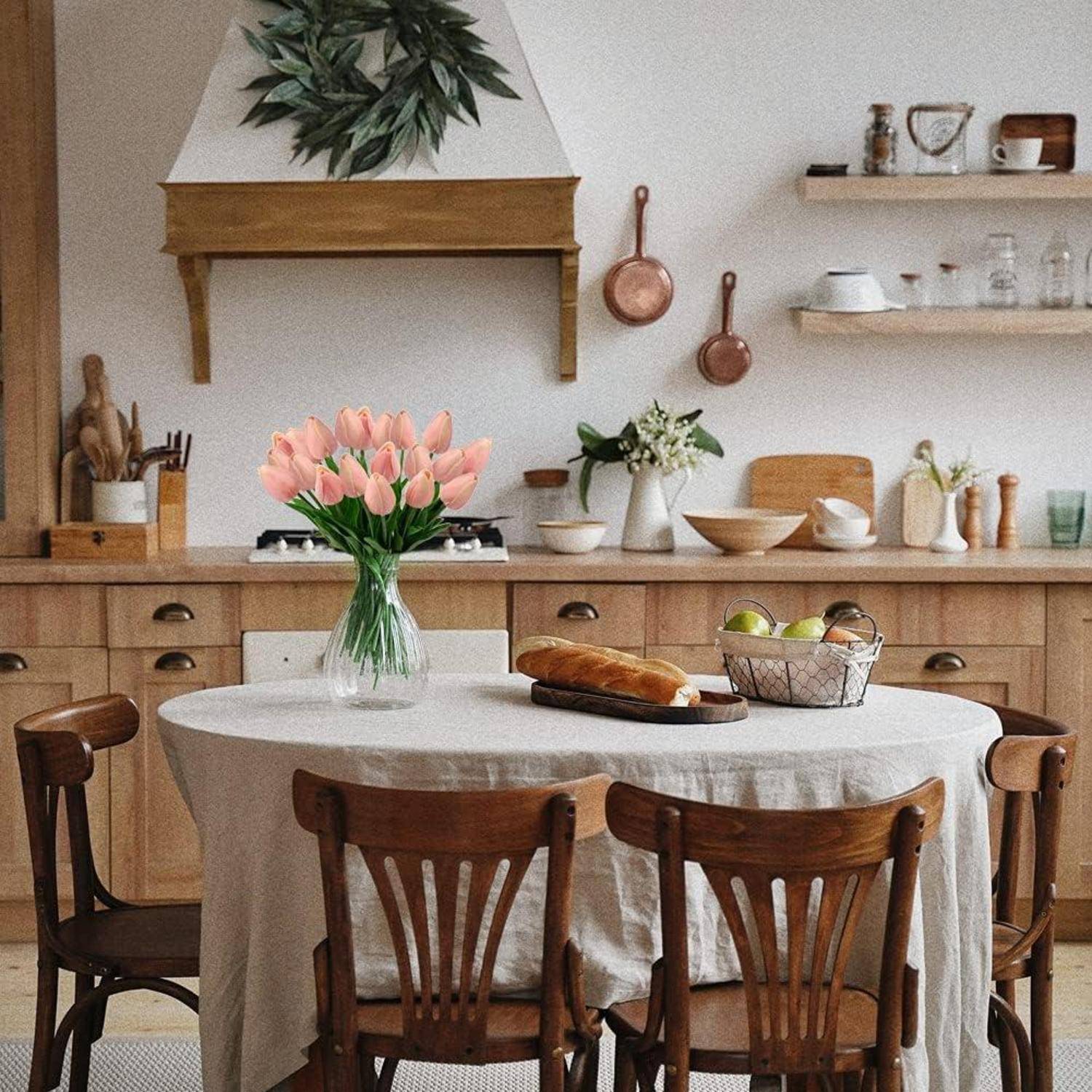 Eine gemütliche Küche zeigt einen Holz-Esstisch mit einer runden Tischdecke, gekrönt von einer Vase mit pinken Tulpen und einem Laib Brot.