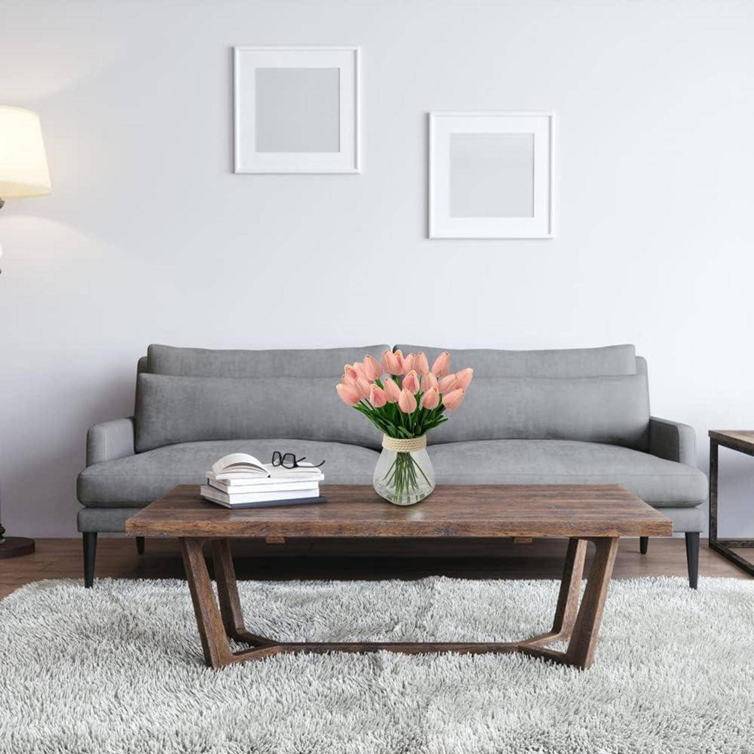 Ein modernes Wohnzimmer mit einem grauen Sofa, einem Holzkaffeetisch und rosa Tulpen in einer Vase. Gerahmte Kunst und eine Lampe befinden sich im Hintergrund.