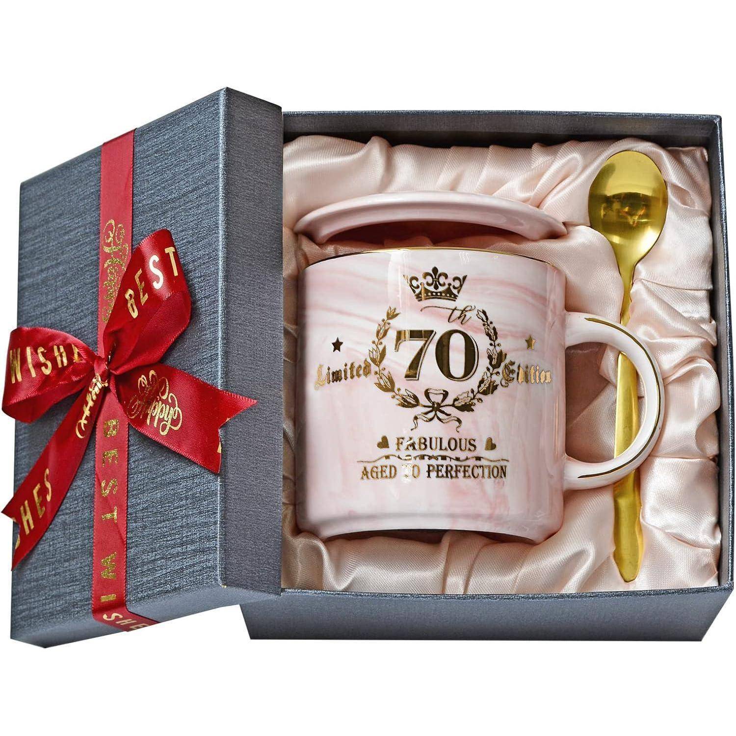 Eine Geschenkbox, die eine Keramiktasse mit der Aufschrift '70' und 'Fabelhaft', einem kleinen Deckel und einem goldenen Löffel enthält, alles auf einem Satin-Futter platziert.