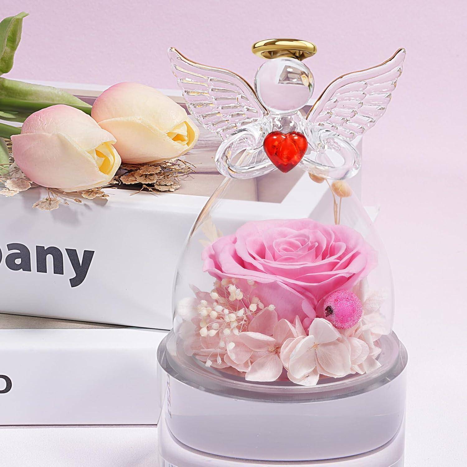 Eine Glasengelfigur mit einem roten Herzen und Flügeln, die eine konservierte rosa Rose und kleine Blumen enthält, wird vor einem weichen rosa Hintergrund präsentiert.
