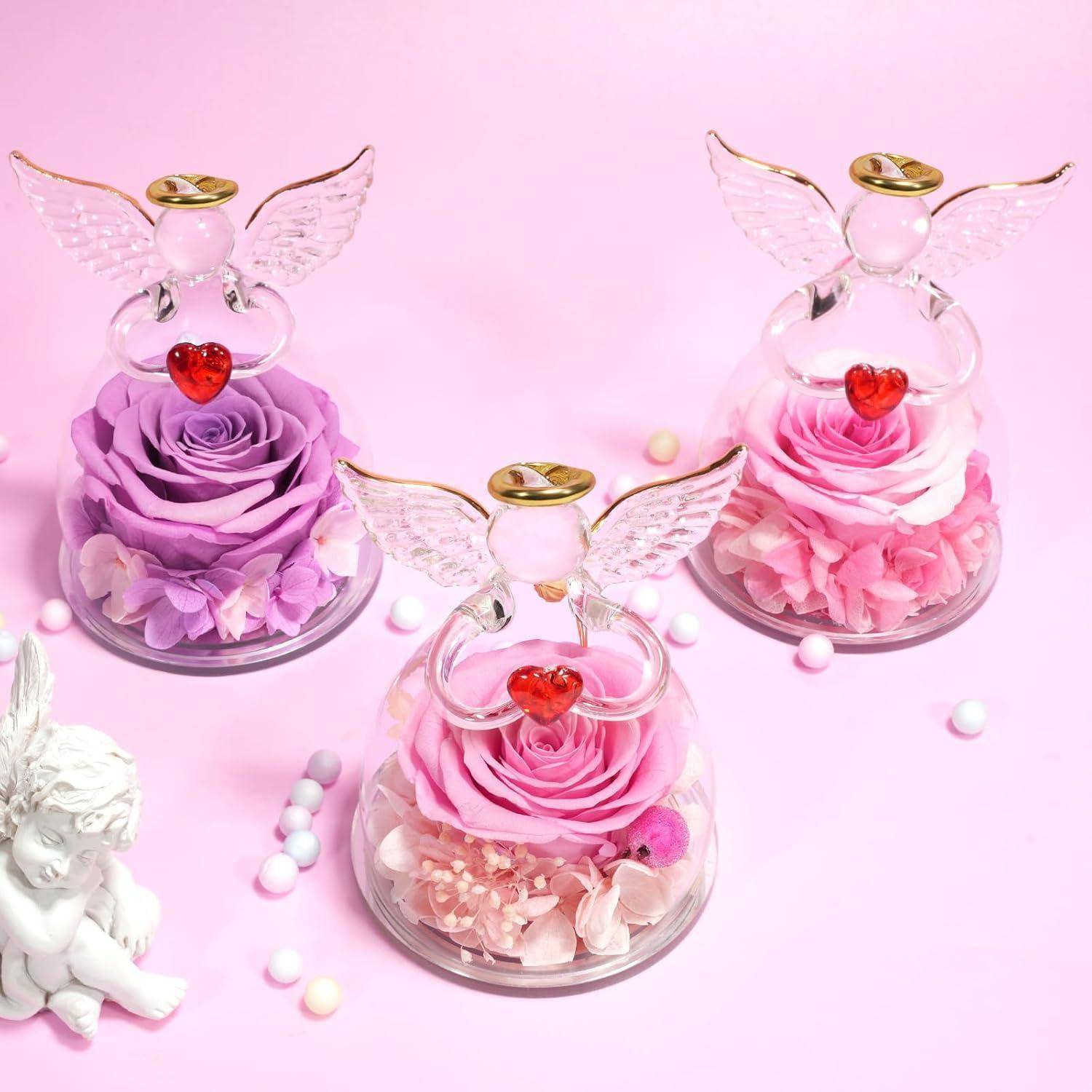 Drei Glasengelfiguren mit Herzakzenten sitzen auf konservierten Rosen in Rosa- und Violetttönen. Eine kleine Engelstatue und pastellfarbene Perlen befinden sich in der Nähe.