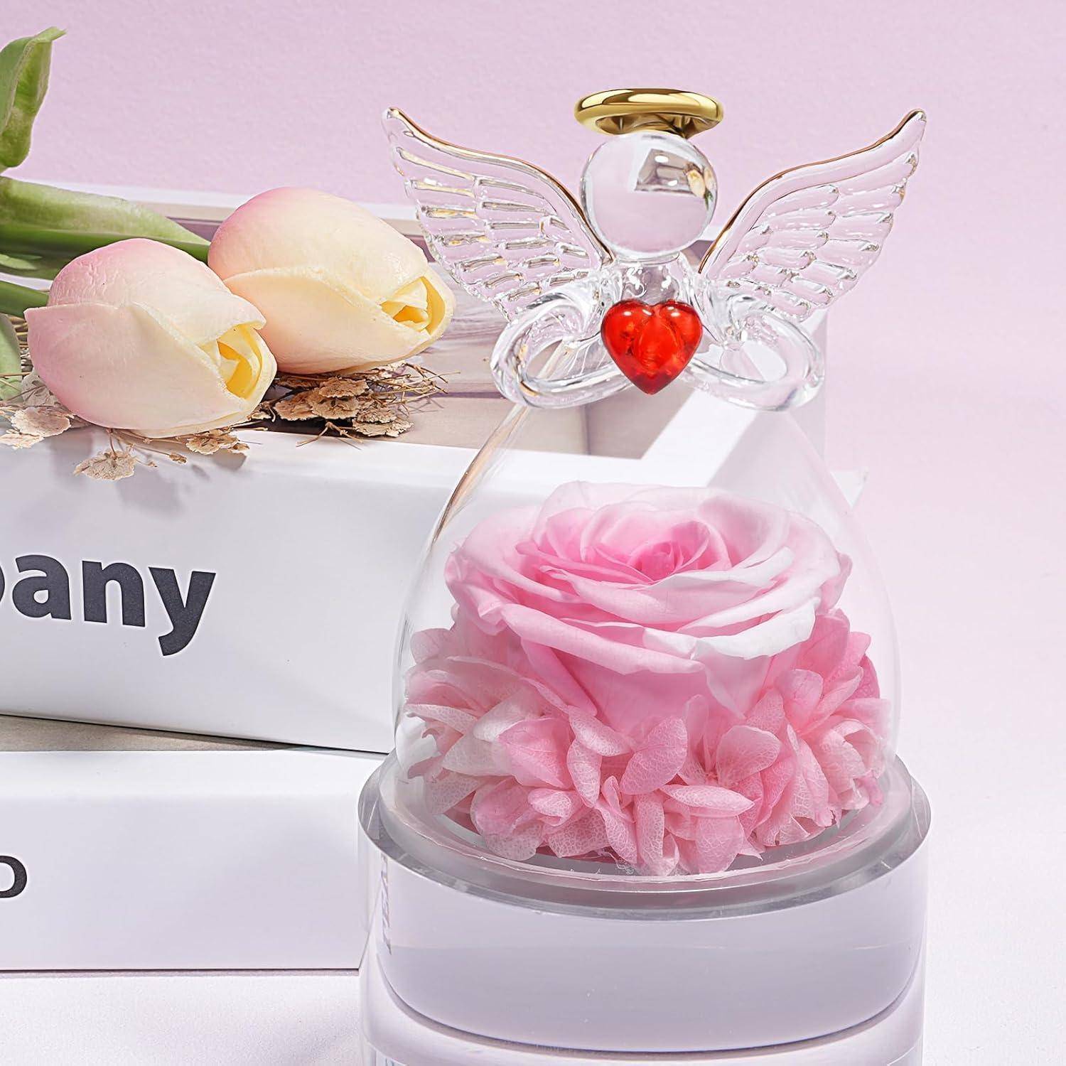 Eine rosa Rose, die in einer Glaskuppel eingeschlossen ist, mit Engelsflügeln und einem roten Herz darüber, flankiert von zwei blassen gelben Rosen auf einem weißen Schild.