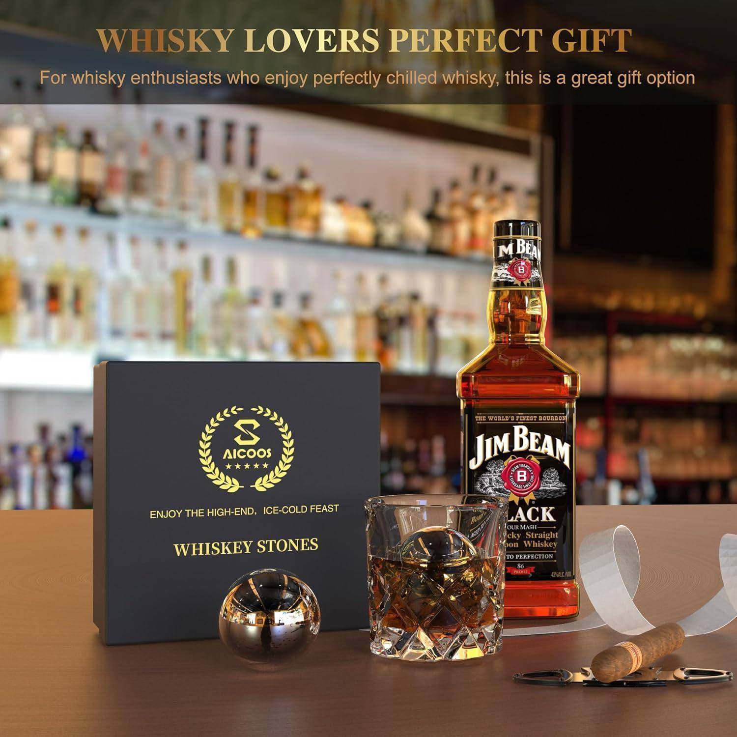 Eine Flasche Jim Beam Whiskey neben einem Glas mit Eissteinen und einer Whiskey-Steine-Box, platziert auf einer Bartheke mit unscharfem Hintergrund.