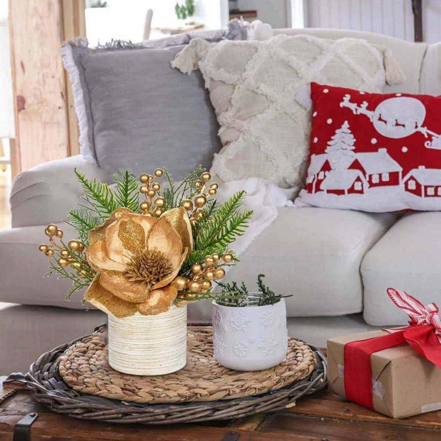 Ein gemütliches Wohnzimmer zeigt eine weiße Couch mit dekorativen Kissen, ein festliches rotes Kissen mit einer Winterszene, eine goldene Blumenarrangement und ein kleines verpacktes Geschenk auf einem Flechttablett.