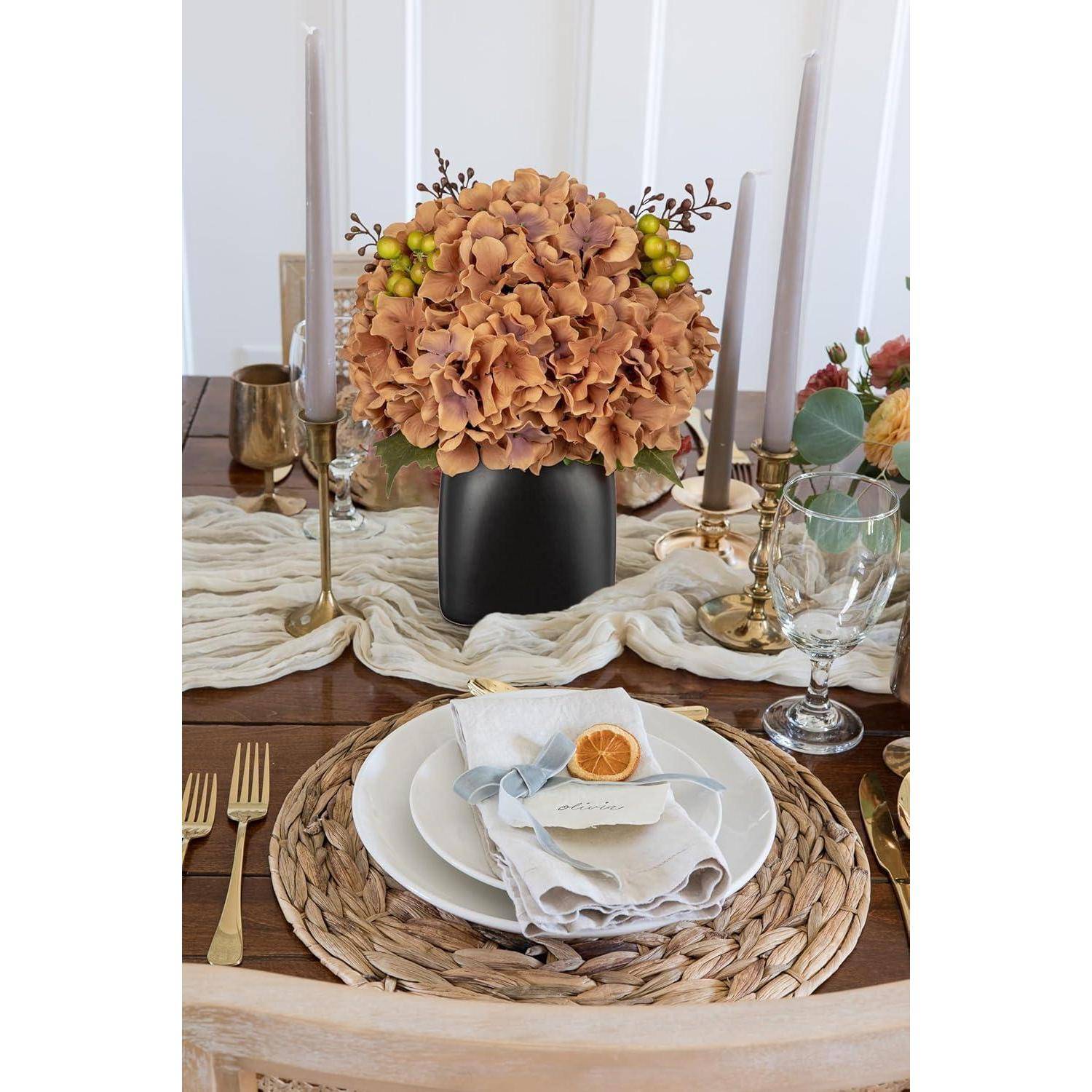 Ein Esstisch, gedeckt mit einem geflochtenen Tischset, weißem Teller, Serviette mit einer getrockneten Orangenscheibe, Weinglas, Besteck, Kerzen und einem Mittelstück aus orangefarbenen Hortensien in einer schwarzen Vase.
