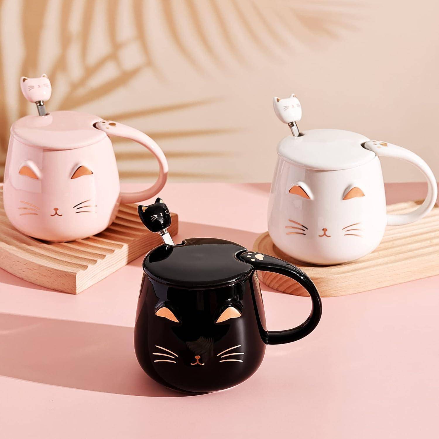 Drei Katzen-Becher mit Katzenkopf-Designs und katzenförmigen Deckeln und Löffeln in Rosa, Weiß und Schwarz, die auf einer rosa Oberfläche platziert sind.