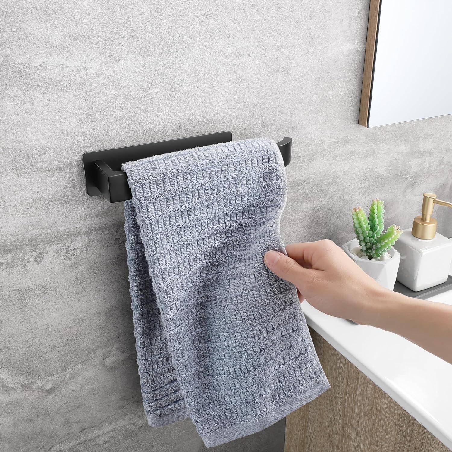 Eine Hand hängt ein graues strukturiertes Handtuch an eine schwarze Wandhalterung neben einem weißen Waschbecken mit einer kleinen Pflanze und einem Seifenspender.