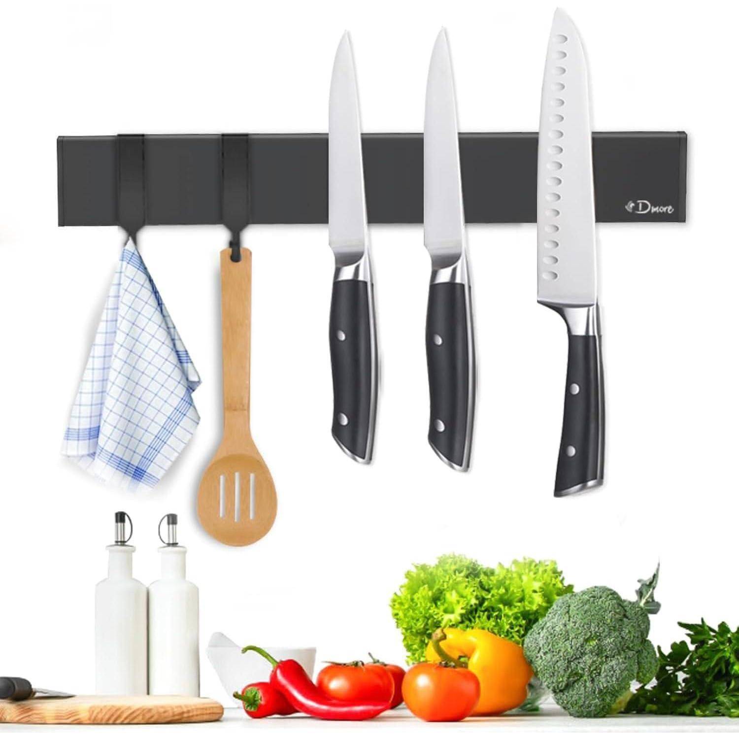 Ein magnetischer Messerhalter an der Wand hält drei Messer, ein Tuch und einen Holzlöffel. Darunter befinden sich Gemüse und Gewürze auf der Arbeitsplatte.