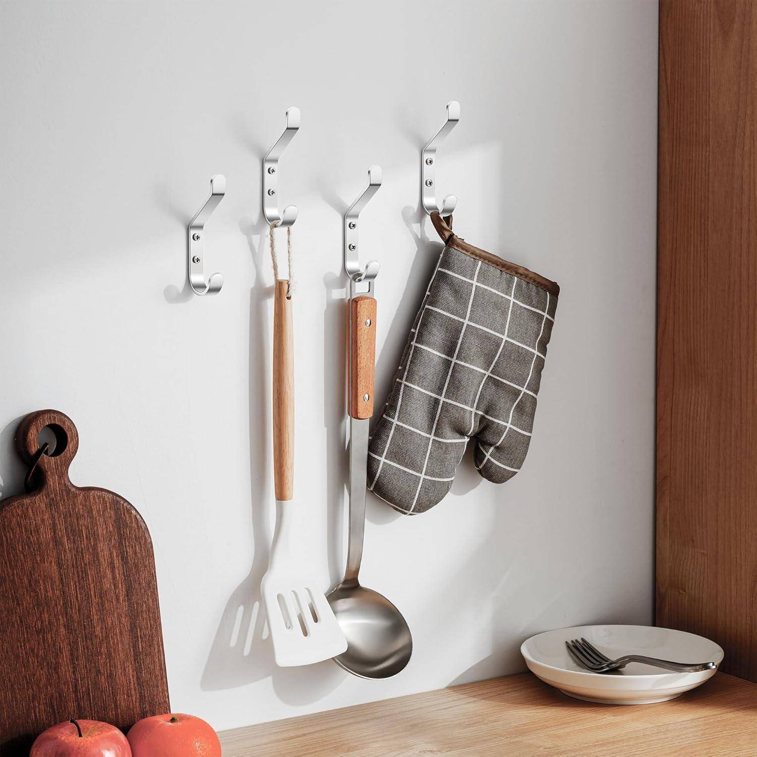 Eine Küchenwand mit Haken, an denen Kochutensilien hängen: ein Pfannenwender, ein Schöpflöffel und ein Ofenhandschuh. Ein Holzschneidebrett, ein Apfel und ein Teller liegen in der Nähe.