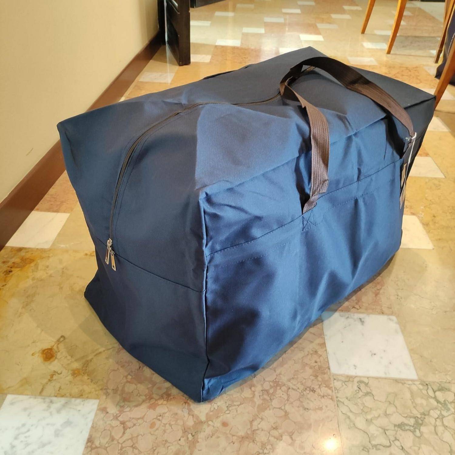 Eine große, blaue, mit Reißverschluss versehene Aufbewahrungstasche mit Griffen steht auf einem gefliesten Boden. Die Tasche ist gefüllt, was sie leicht ausbauchen lässt.