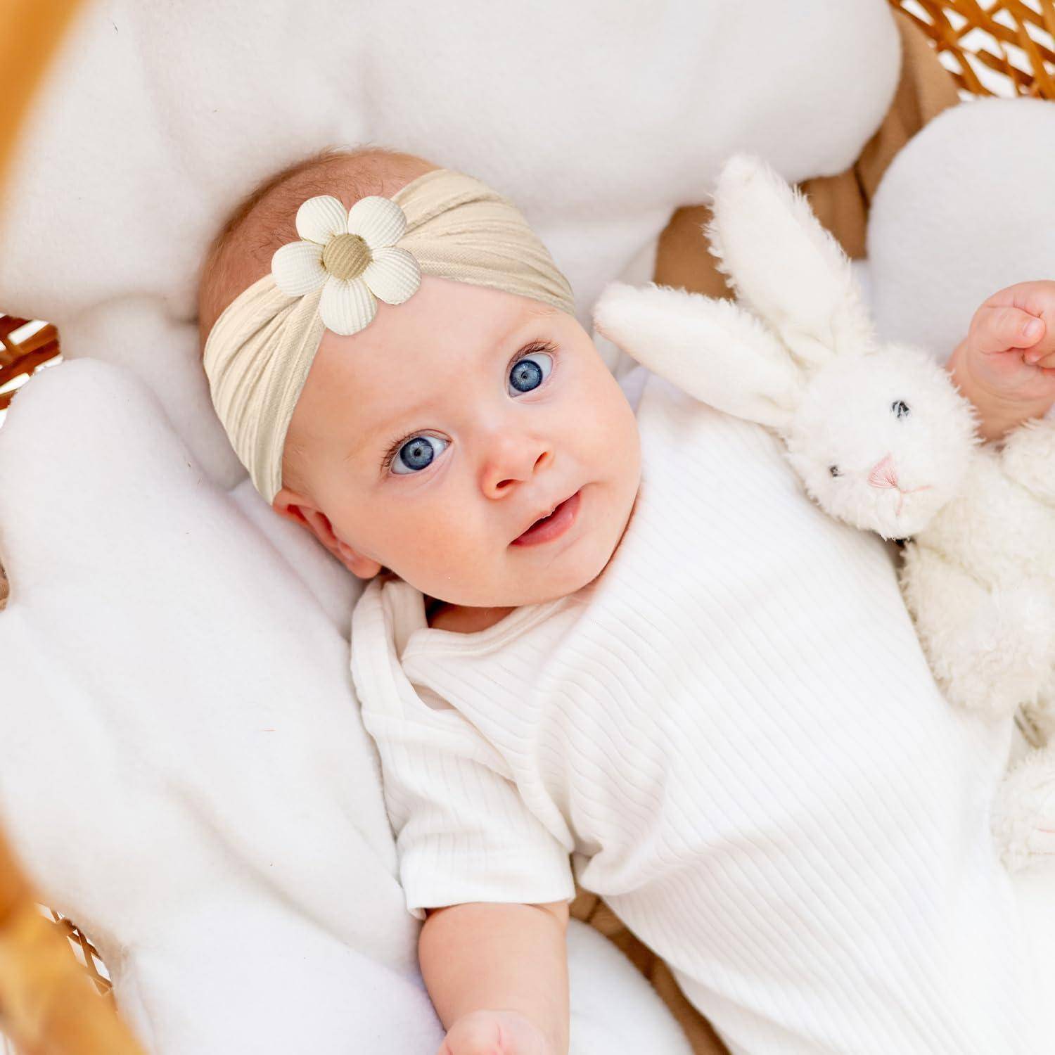 Ein Baby mit blauen Augen und einem beigen Stirnband liegt in einer weißen Decke und hält einen weichen, weißen Stoffhasen.