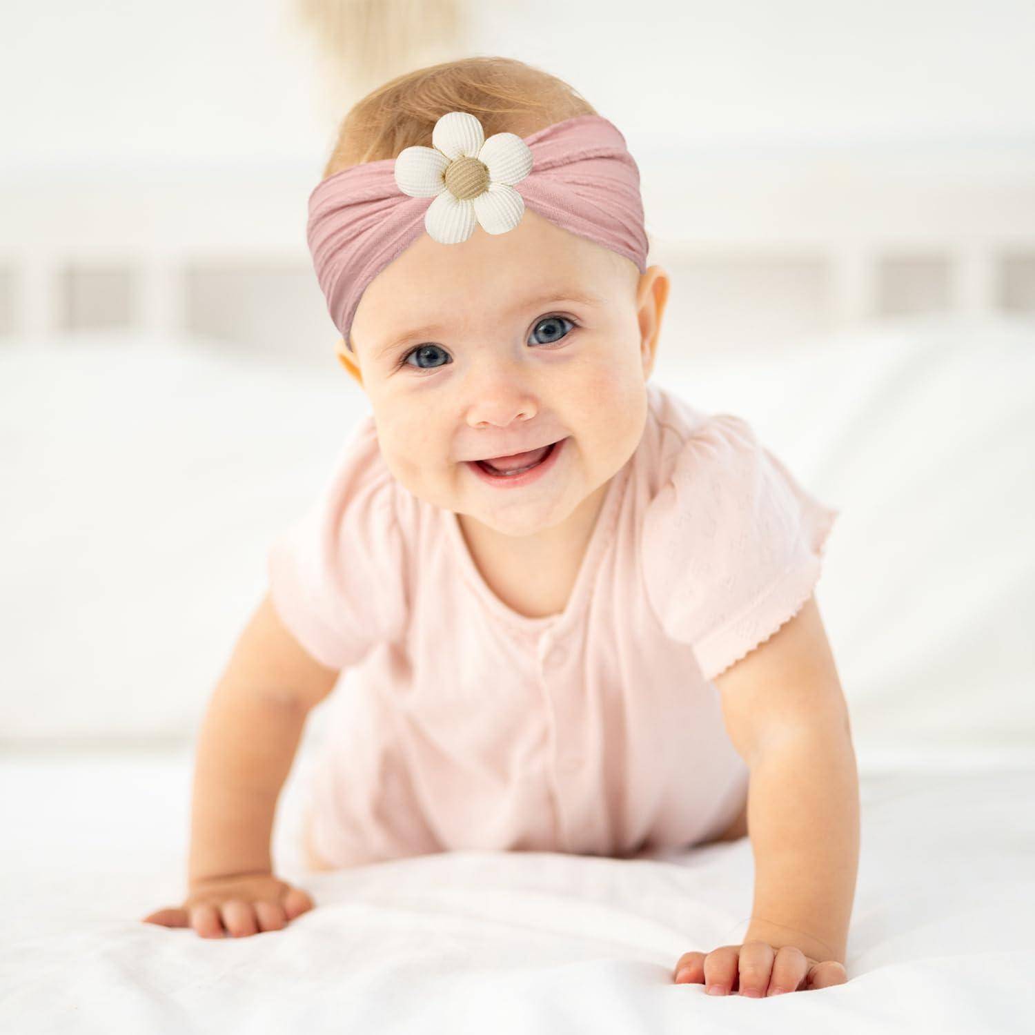 Ein lachendes Baby mit einem rosa Haarband mit einer Blume sitzt auf einem weißen Bett. Das Baby trägt einen rosa Anzug.