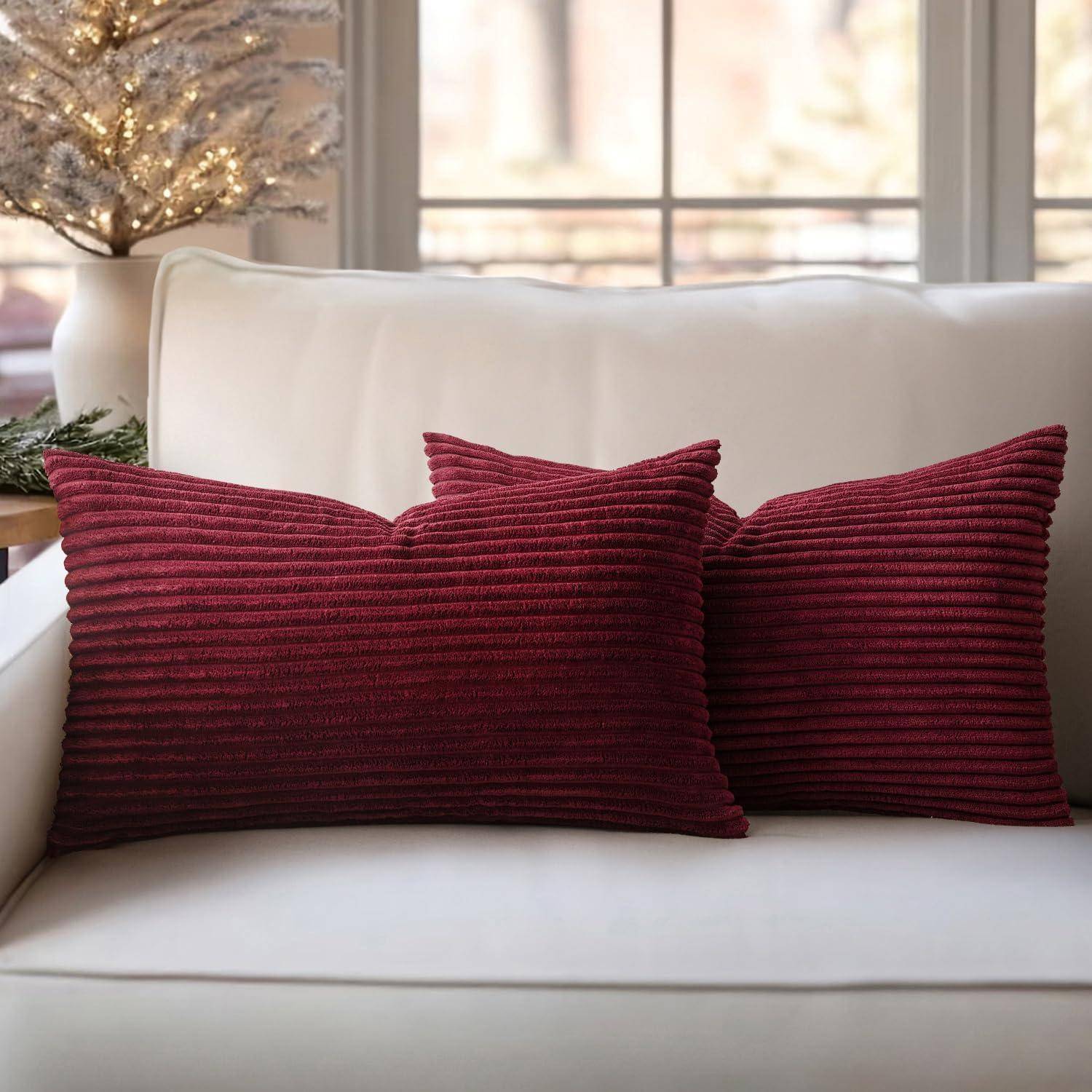 Ein weißes Sofa mit zwei burgunderroten gerippten Kissen steht in einem gemütlichen Raum mit einem beleuchteten, dekorativen Baum und großen Fenstern im Hintergrund.