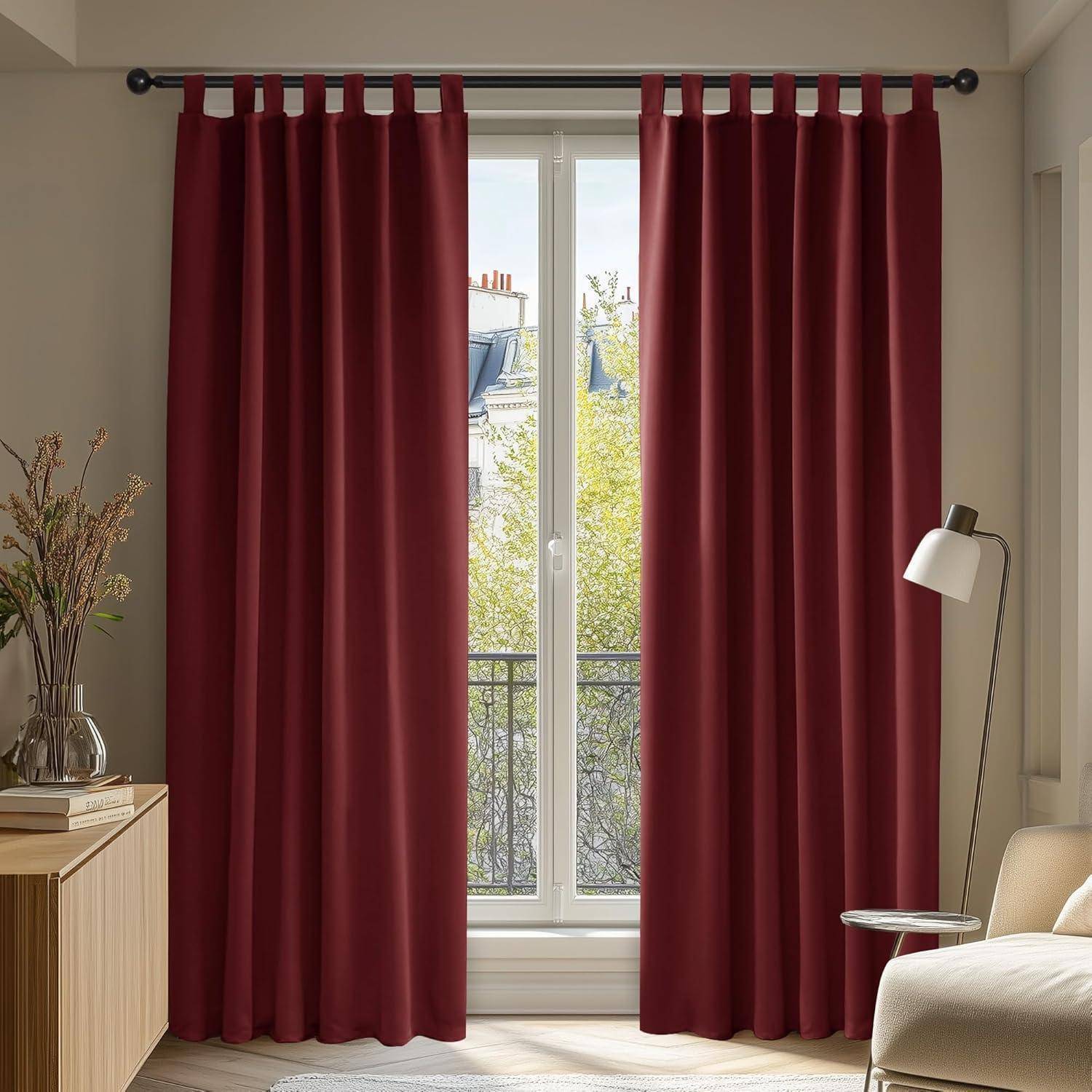 Rote Vorhänge hängen über einem Fenster und geben den Blick auf Bäume und Dächer draußen frei. Eine moderne Lampe steht neben einem Holzschrank.