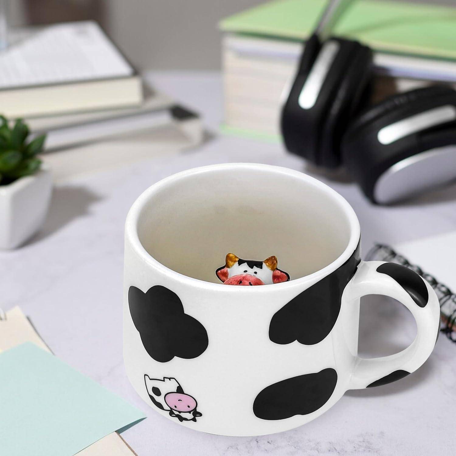Eine weiße Keramiktasse mit schwarzen Flecken, die eine Kuh ähnelt, mit einer kleinen Kuhfigur, die von innen herausschaut. Sie steht auf einem Schreibtisch in der Nähe von Kopfhörern und Büchern.