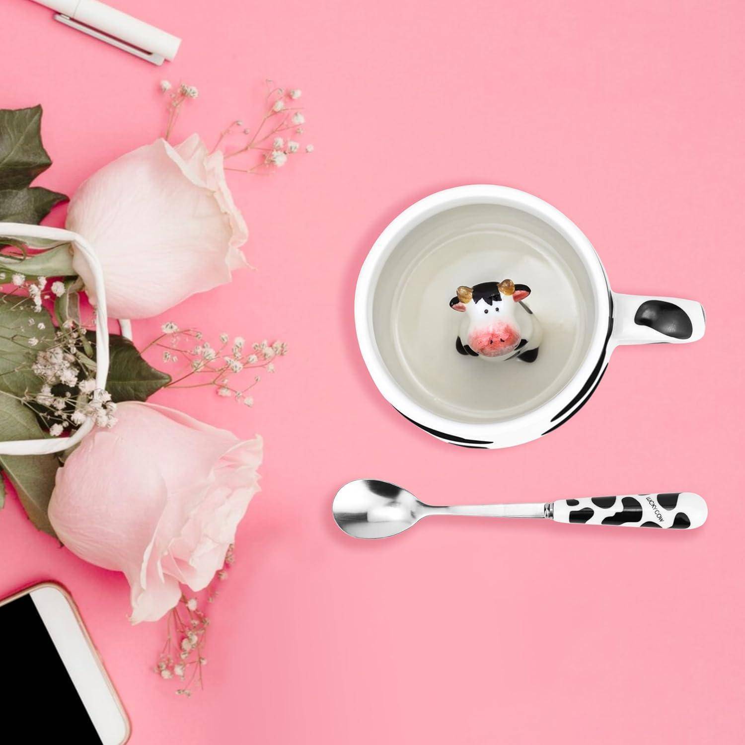 Luftaufnahme einer Teetasse mit Kuhdesign im Inneren, neben einem passenden Löffel. In der Nähe befinden sich rosa Rosen und ein Telefon auf einer rosa Oberfläche.