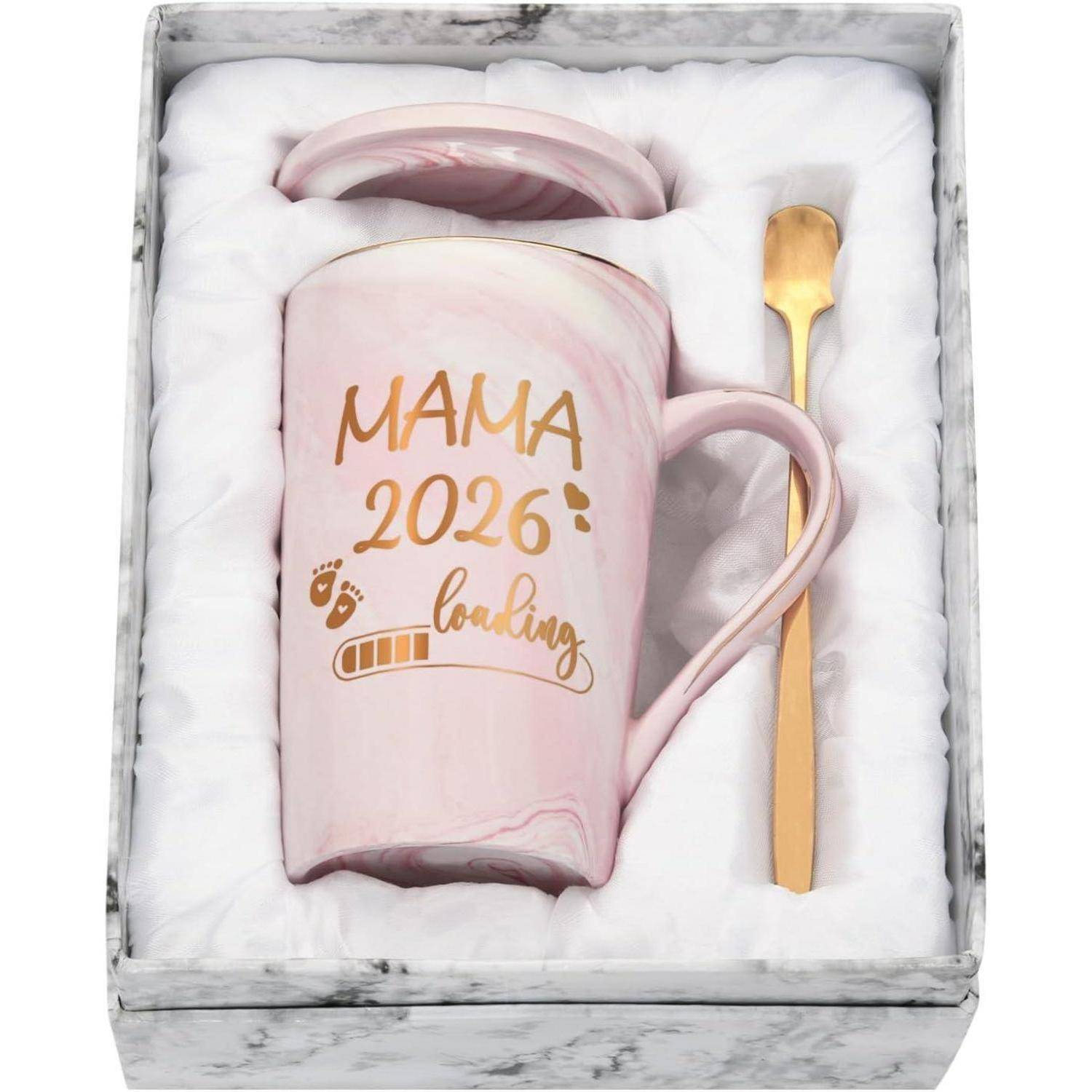 Eine rosa Tasse mit 'MAMA 2026 wird geladen' in Gold, versehen mit Fußspuren, geliefert mit einem Deckel und einem goldenen Löffel in einer gepolsterten Geschenkbox.