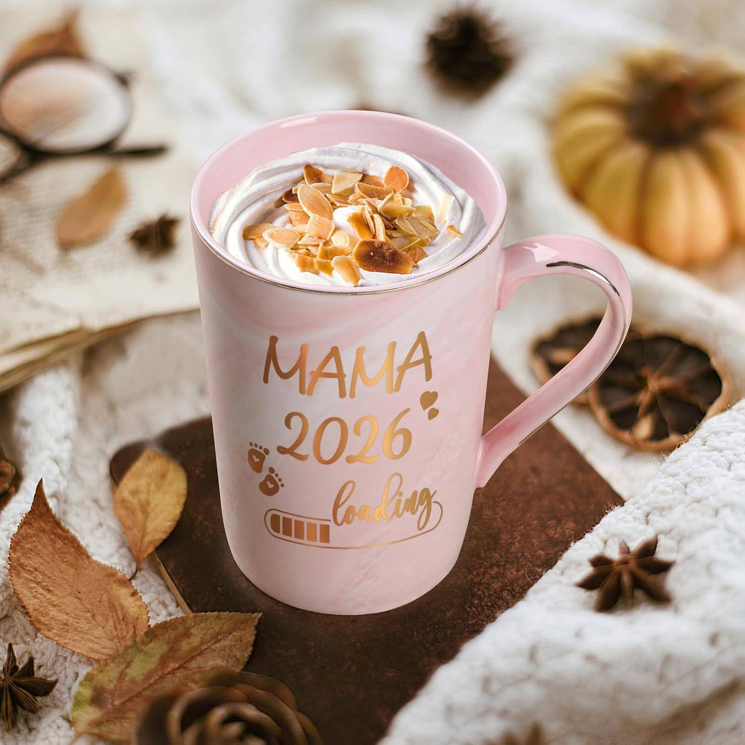 Eine pinke Tasse mit der Aufschrift 'MAMA 2026 lädt', gefüllt mit Schlagsahne und Mandeln. Der Hintergrund zeigt Herbstblätter und einen kleinen Kürbis.