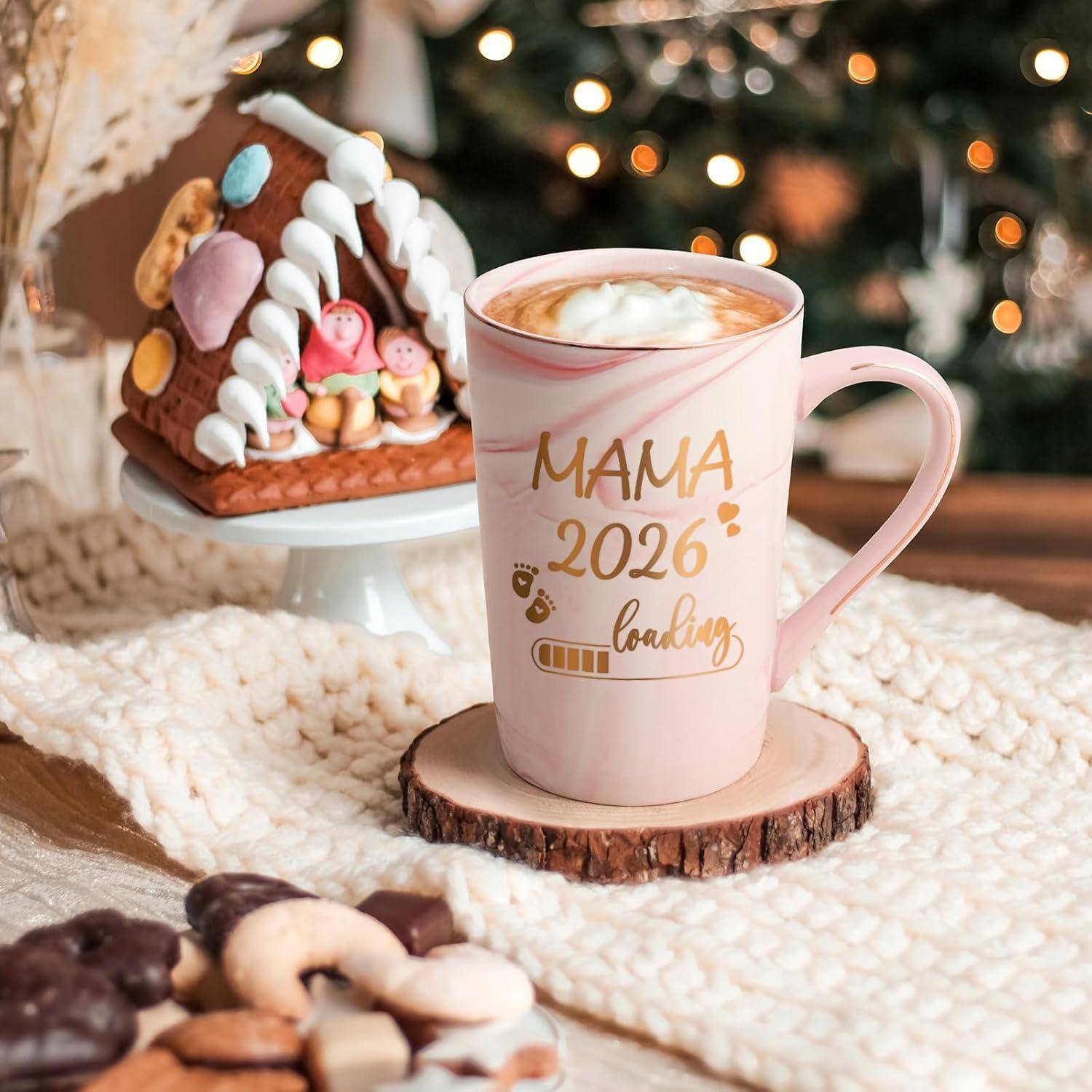 Eine Tasse mit der Aufschrift 'MAMA 2024 lädt' steht auf einem Holzuntersetzer, umgeben von Keksen und einem Lebkuchenhaus, in einer festlichen Umgebung mit einem Weihnachtsbaum.