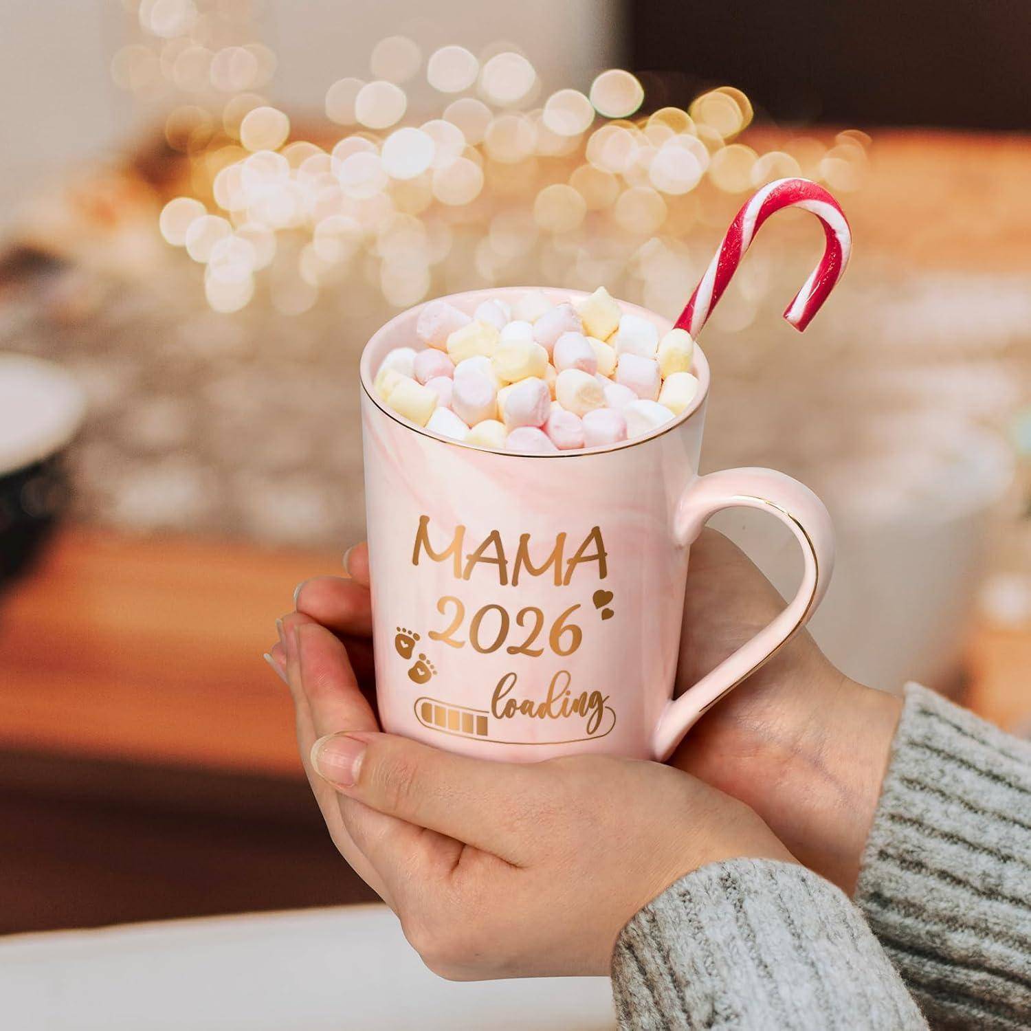Eine Hand hält eine Tasse, gefüllt mit Marshmallows und Zuckerstangen. Auf der Tasse steht „MAMA 2026 lädt