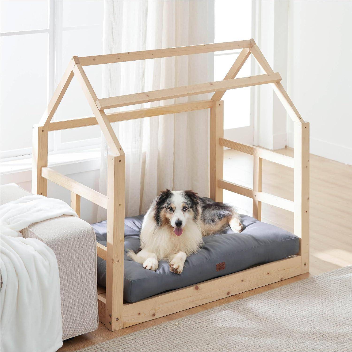 Ein Hund liegt auf einem grauen Kissen in einem holzförmigen, hausähnlichen Hundebett neben einem Sofa in einem hellen Raum mit natürlichem Licht.