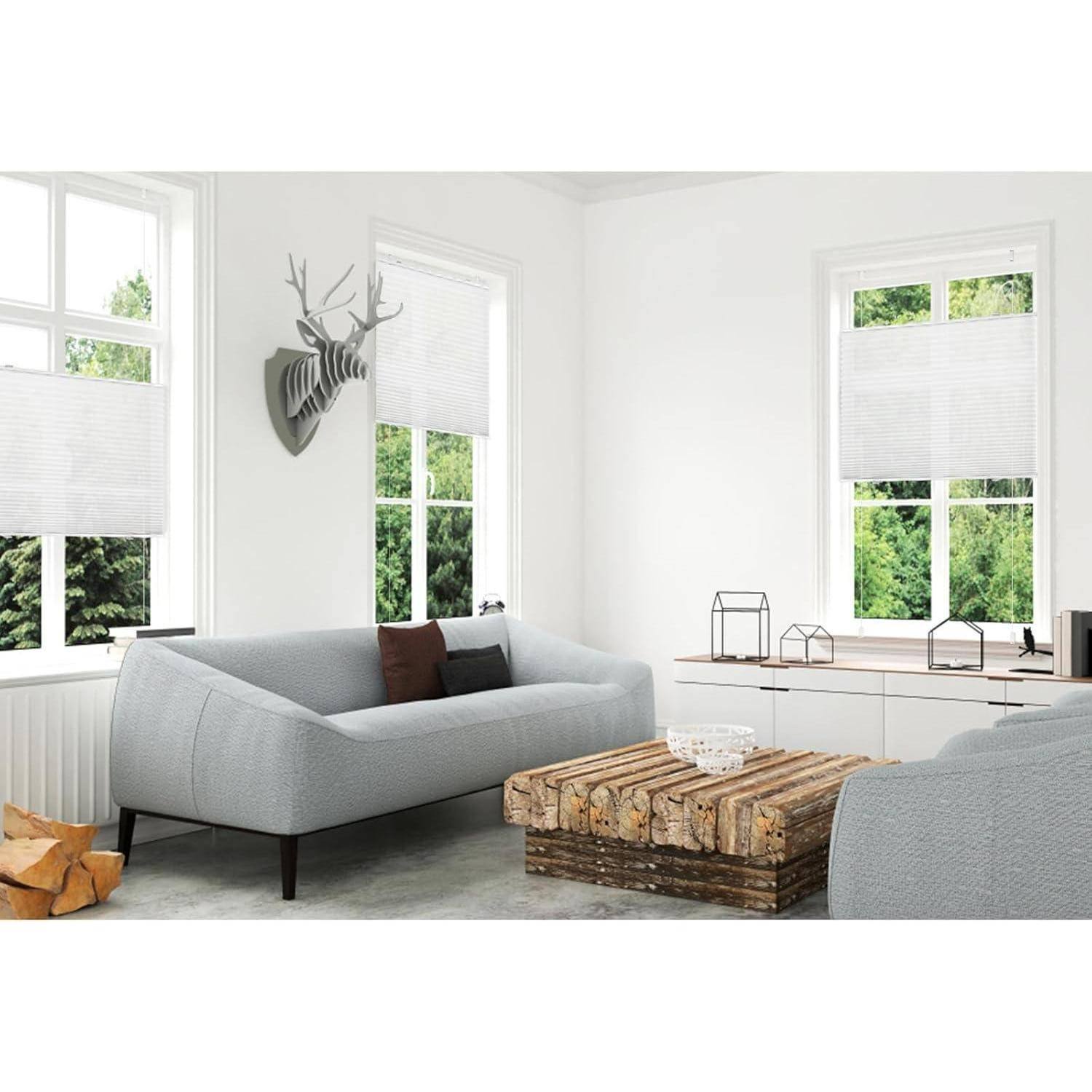 Ein modernes Wohnzimmer mit einem grauen Sofa, einem Couchtisch aus Holz und weißen Wänden. Große Fenster lassen natürliches Licht herein. Ein Hirschkopf-Deko hängt an der Wand.