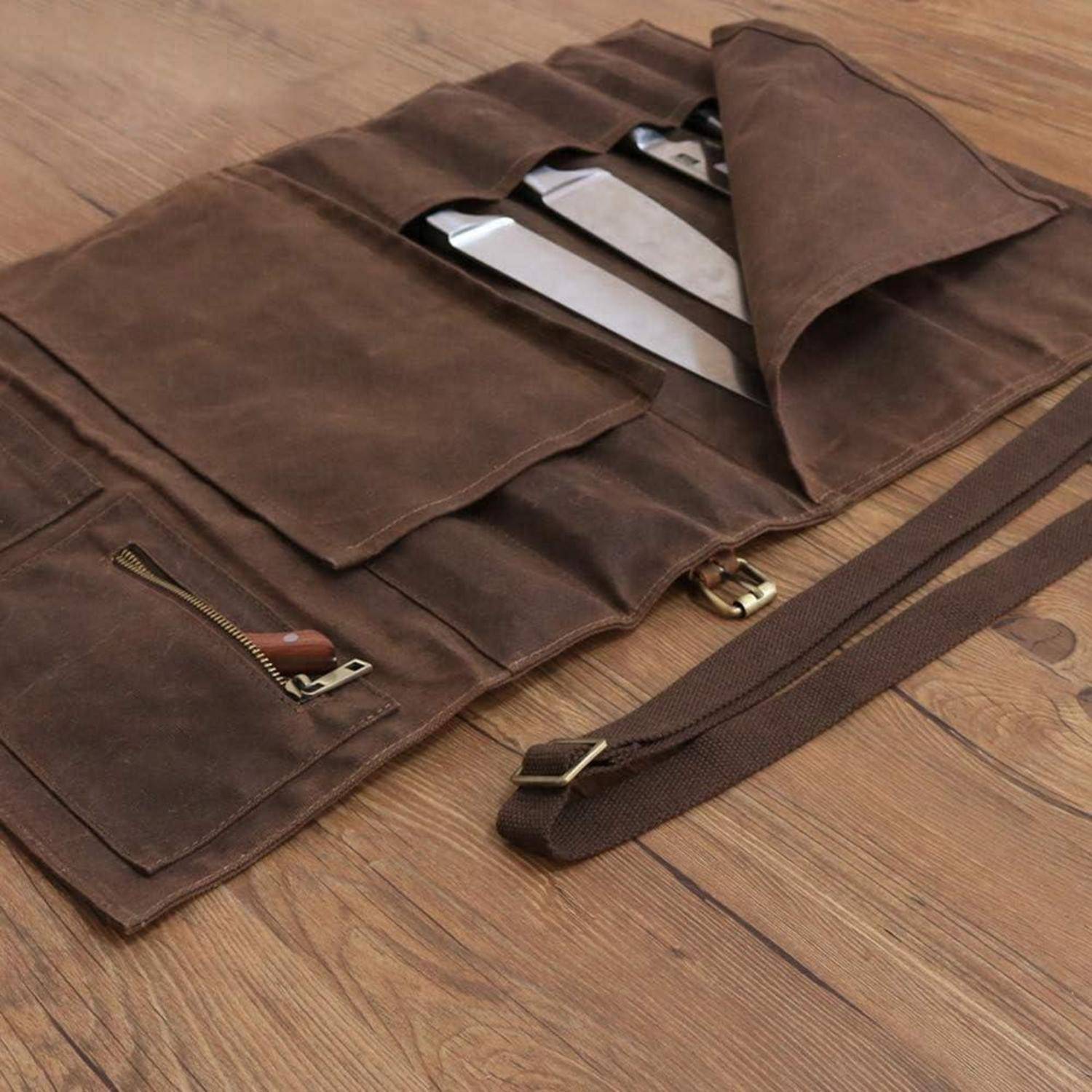 Eine braune Ledermessertasche ist auf einem Holzboden geöffnet und zeigt verschiedene Messerfächer und einen Trageriemen.