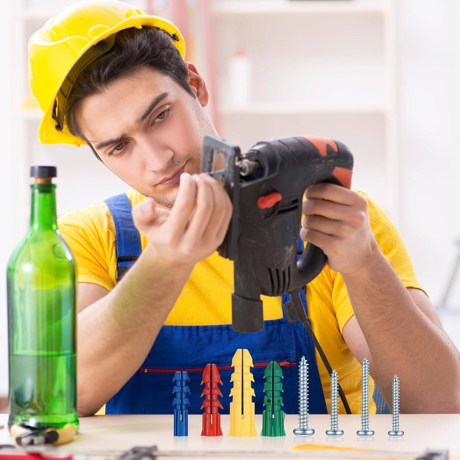 Ein Bauarbeiter mit gelbem Schutzhelm und blauen Overalls untersucht einen Akkuschrauber, während Wanddübel und Schrauben auf dem Tisch liegen.