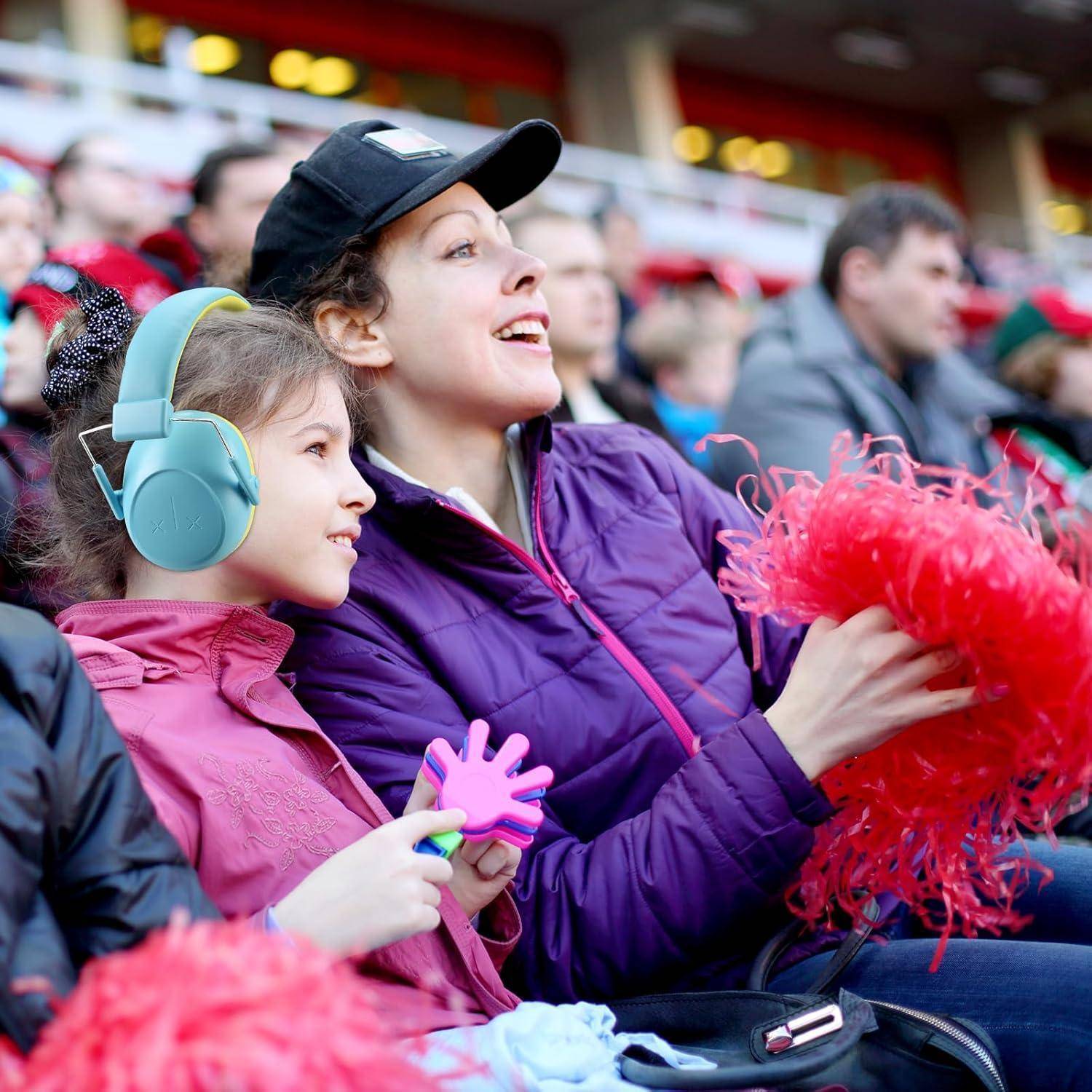 Eine Frau und ein Kind mit Kopfhörern jubeln begeistert bei einer Sportveranstaltung und halten rote Pompons inmitten einer lebhaften Menschenmenge.