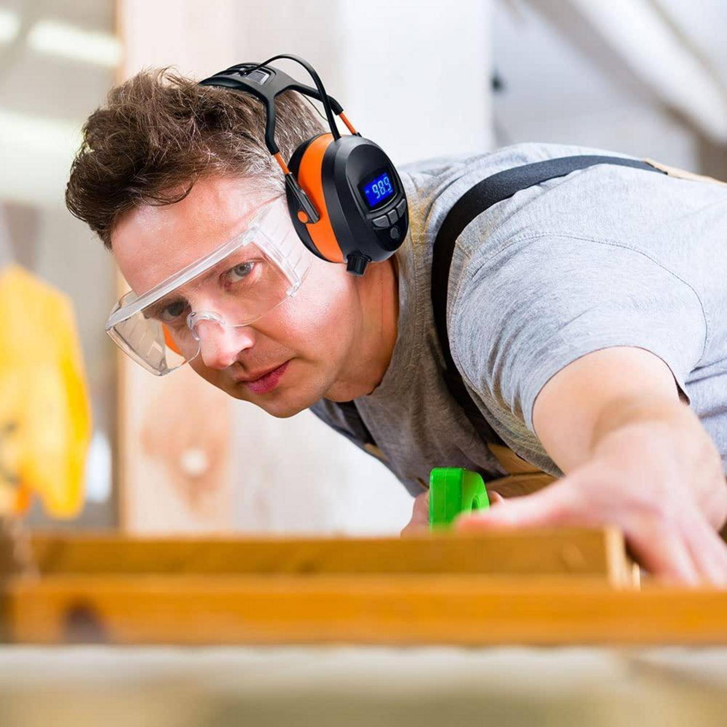 Ein Arbeiter mit Schutzbrille und Gehörschutz ist konzentriert bei der Holzbearbeitung und verwendet ein grünes Werkzeug an einem Holzbrett im Innenraum.