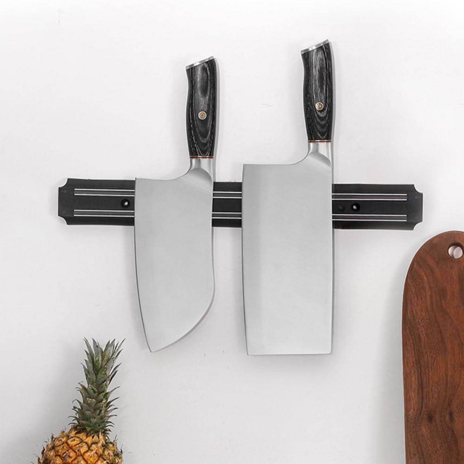 Zwei große Küchenmesser hängen an einem schwarzen Magnetstreifen, der an einer Wand befestigt ist. Darunter liegt eine Ananas, und rechts davon steht ein Holzschneidebrett.