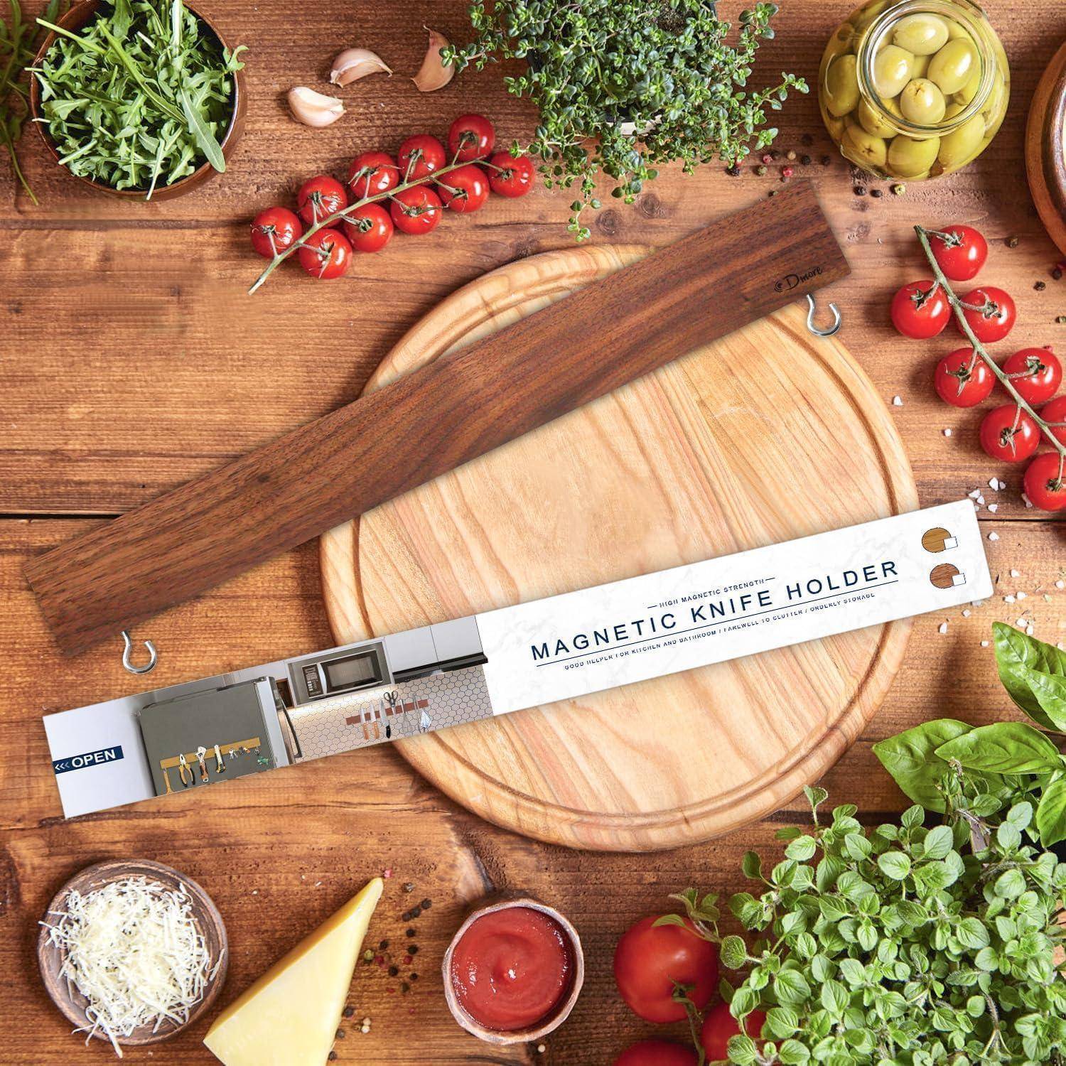 Ein hölzerner magnetischer Messerhalter liegt auf einem Holztisch, umgeben von Kirschtomaten, Kräutern, einer Knoblauchknolle, Oliven und Küchenutensilien.