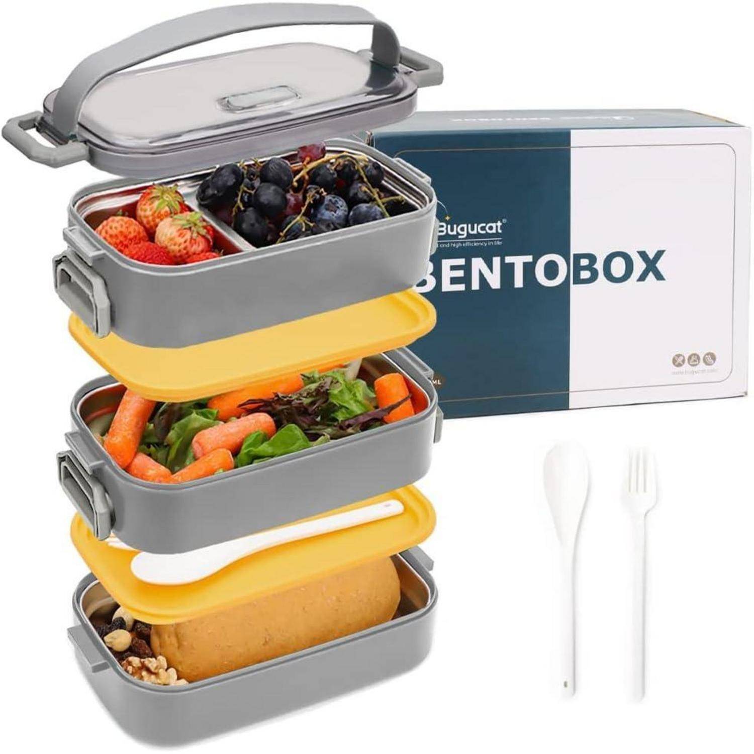 Eine dreistöckige graue Bento-Box mit verschiedenen Lebensmitteln: Erdbeeren, Trauben, Karotten, Salat und Brot. Verpackung und Besteck sind sichtbar.