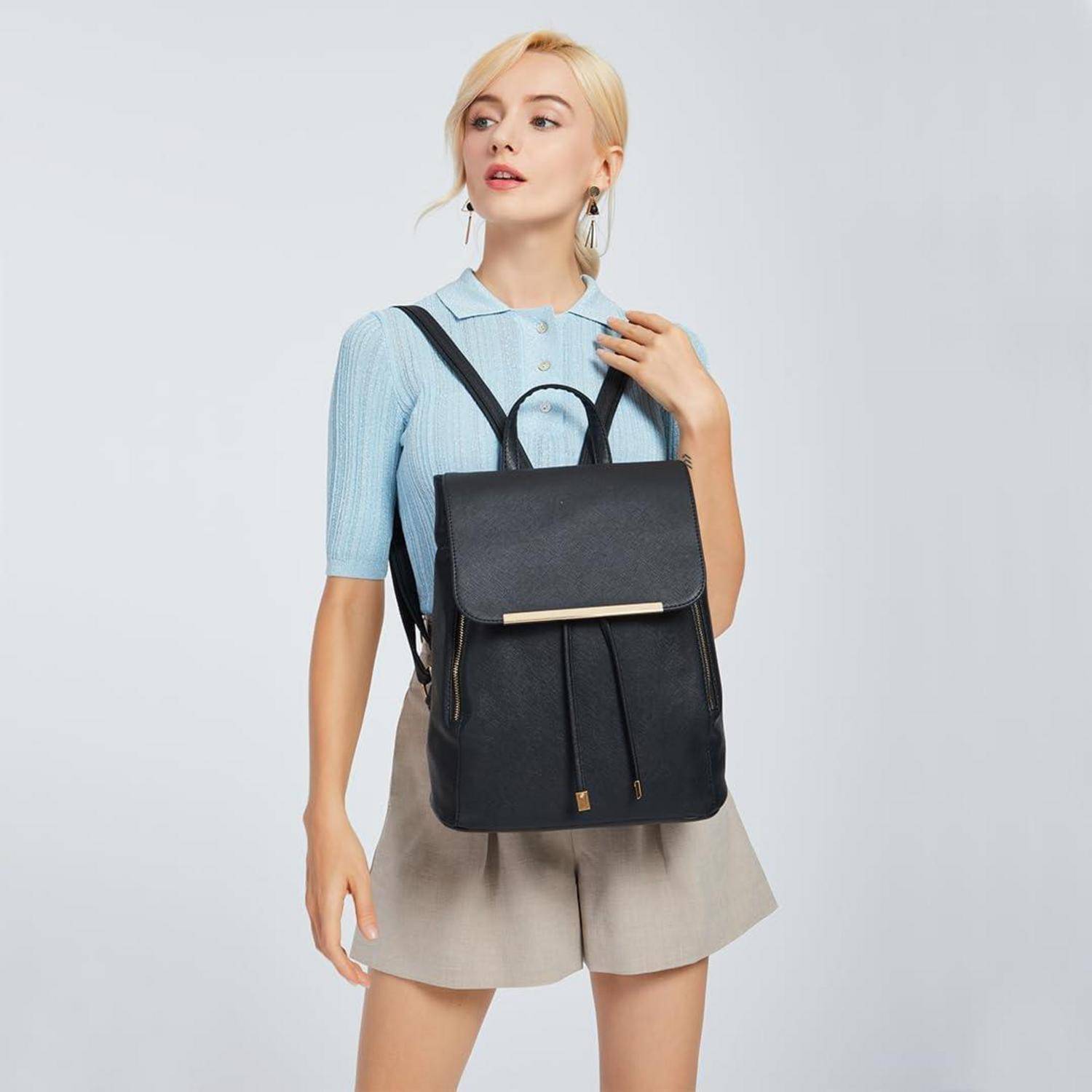 Eine Person mit kurzen blonden Haaren trägt ein blaues Hemd und beige Shorts und hält einen schwarzen Rucksack vor einem hellgrauen Hintergrund.