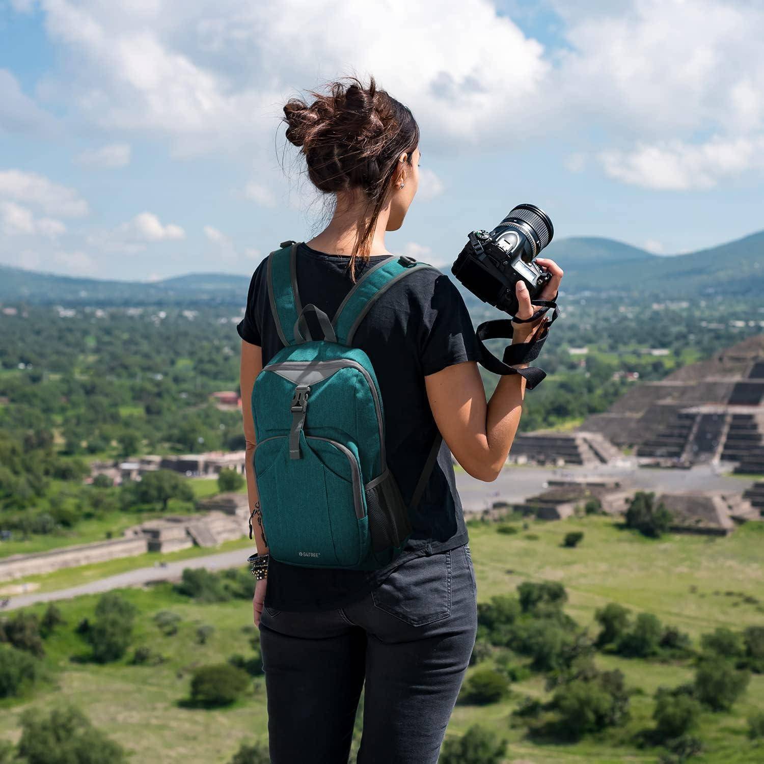 Eine Frau mit einem Rucksack und einer Kamera blickt auf alte Ruinen vor einer bergigen Landschaft unter einem klaren Himmel.