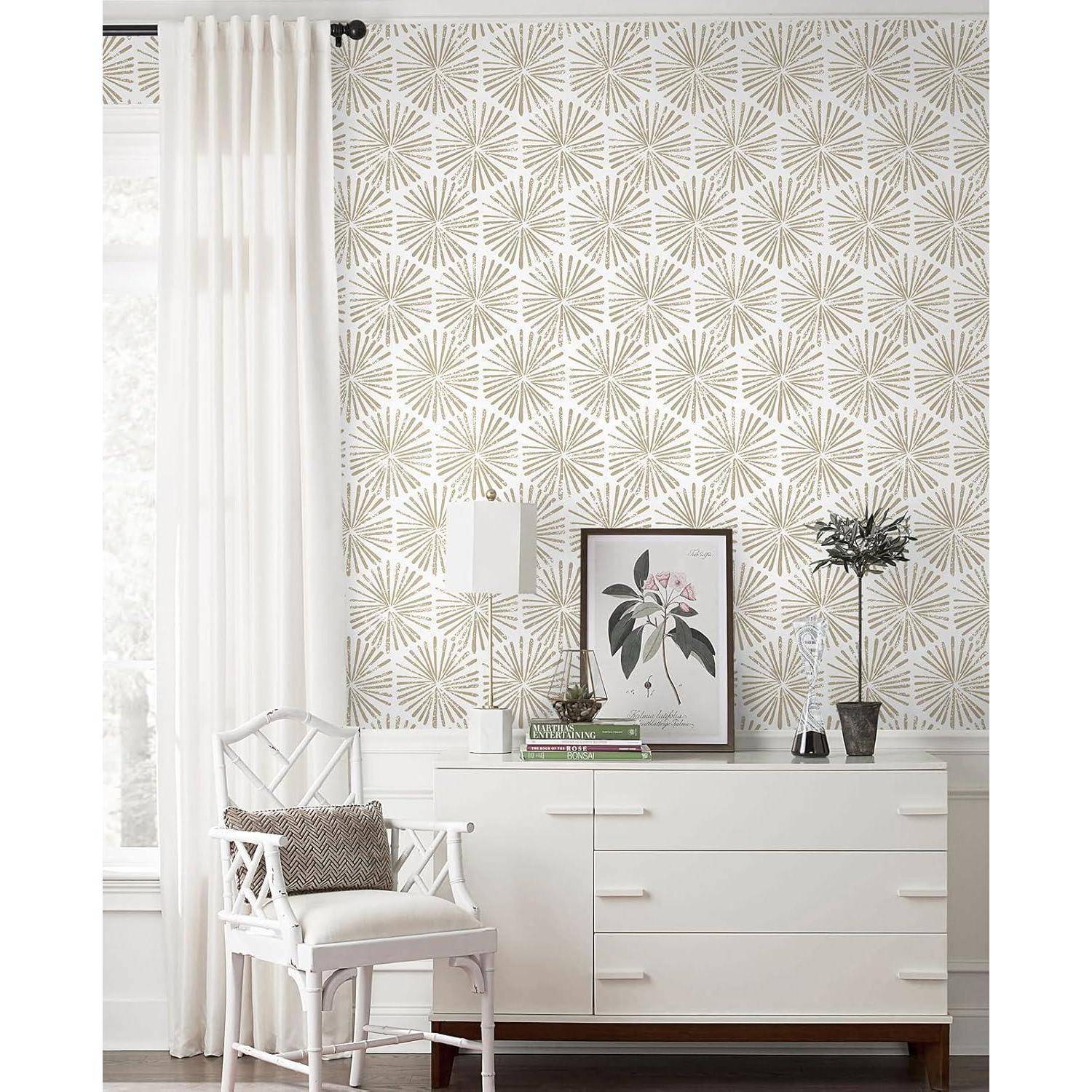 Ein weißer Schrank mit einem Blumenbild, einer kleinen Pflanze und Büchern darauf steht vor einer Wand mit goldenem Sternenmuster. Links davon befinden sich ein weißer Stuhl und ein Vorhang.