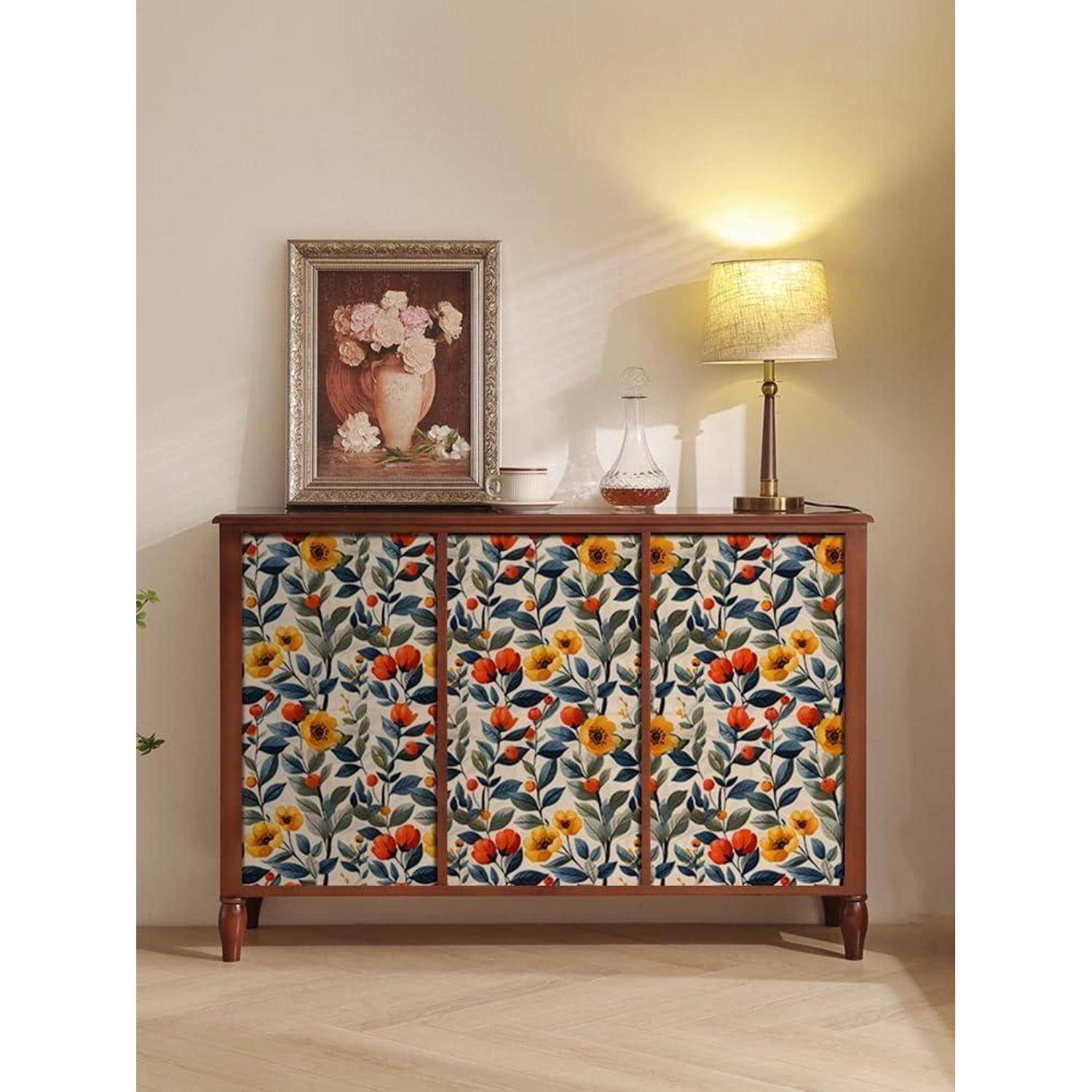 Ein Holzsideboard mit blumenmusterverzierten Türen, einem gerahmten Blumengemälde, einer Glaskaraffe und einer brennenden Lampe auf der Oberfläche, vor einer neutralen Wand.