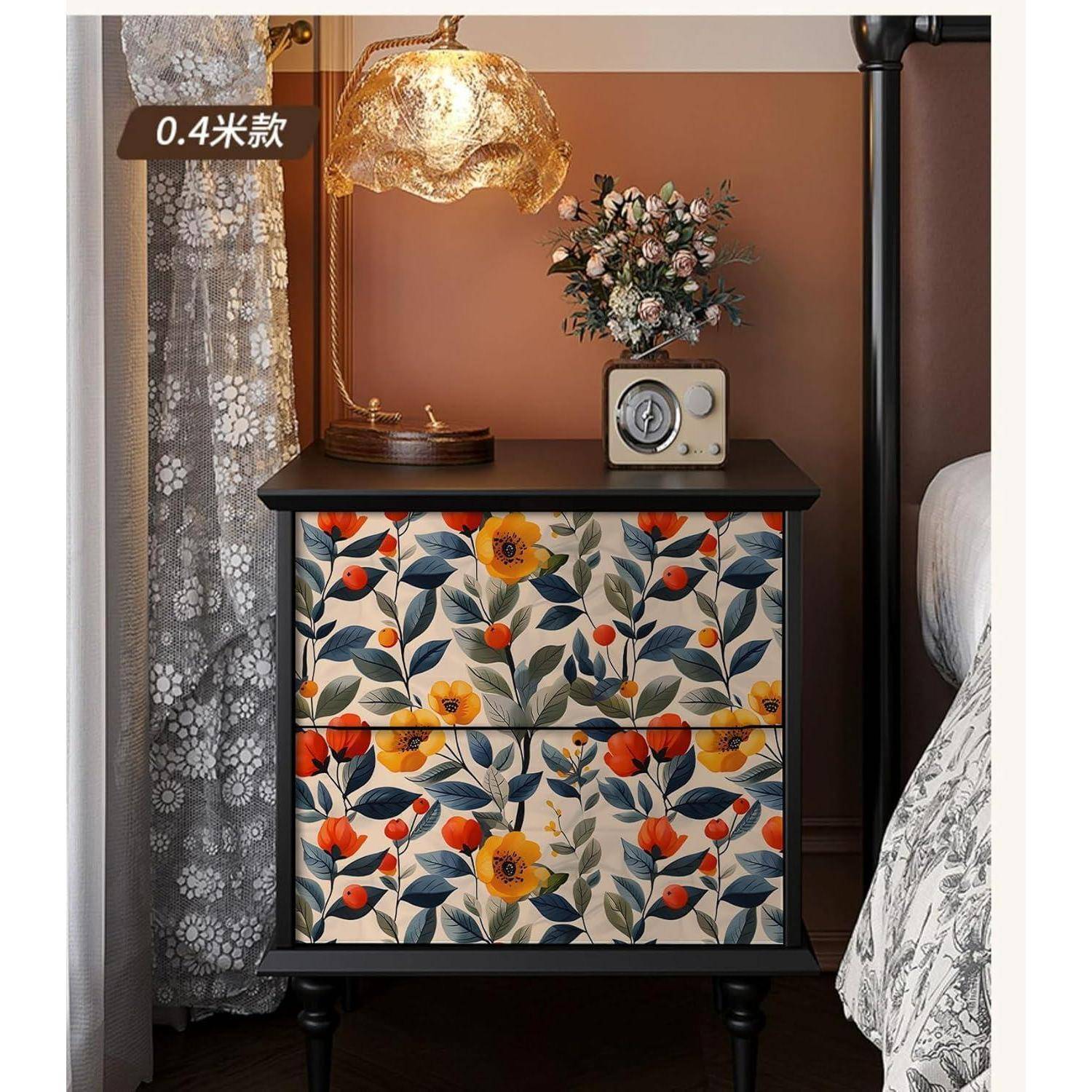Eine kleine Kommode mit zwei Schubladen, verziert mit einem bunten Blumenmuster, steht neben einem Bett. Eine Lampe, Blumen und eine Uhr stehen auf der Oberfläche.