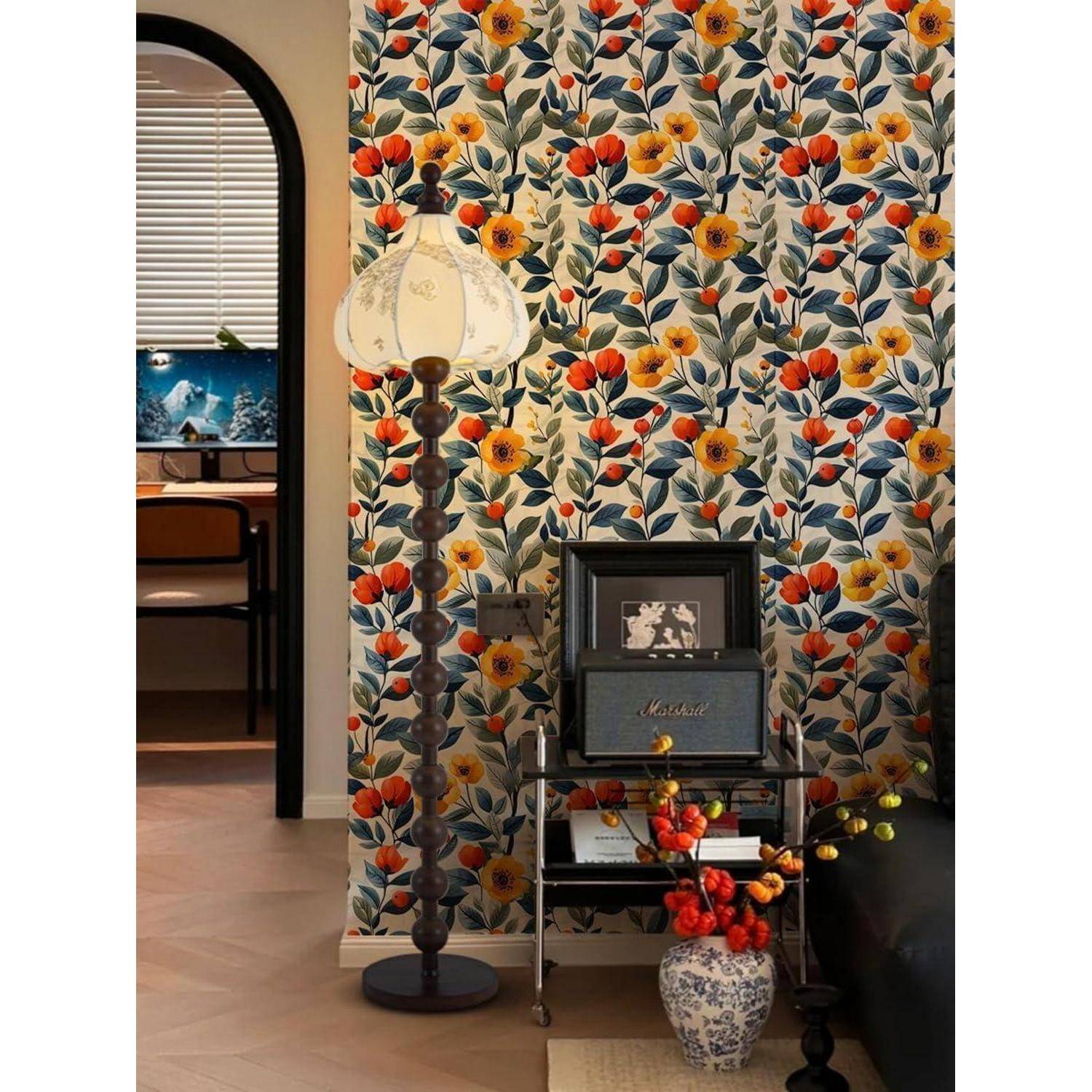 Eine Ecke eines Wohnzimmers mit bunter Blumentapete, einer Vintage-Stehlampe und einem Beistelltisch, auf dem gerahmte Bilder und eine Vase mit Blumen stehen.