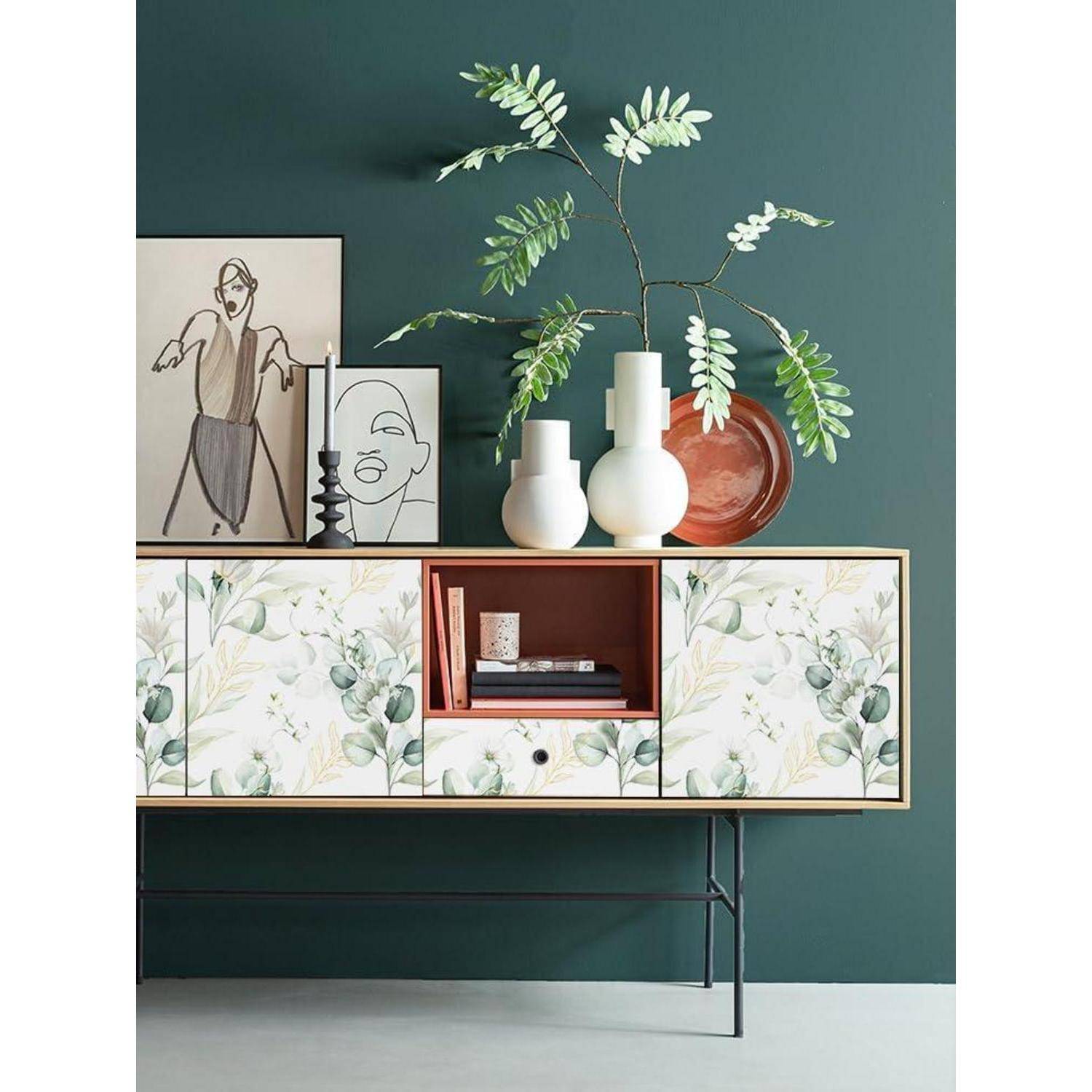 Ein stilvolles Kabinett mit blumenmusterverzierten Türen, auf dem abstrakte Kunst, weiße Vasen, eine rote Schale und ein Zweig mit Blättern vor einer grünen Wand ausgestellt sind.