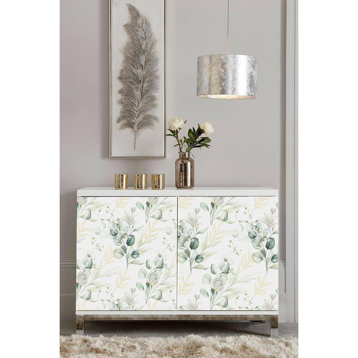 Ein dekorativer Sideboard mit Blumenmuster wird von drei Messingvasen mit weißen Blumen gekrönt und von einer silbernen Pendelleuchte beleuchtet.