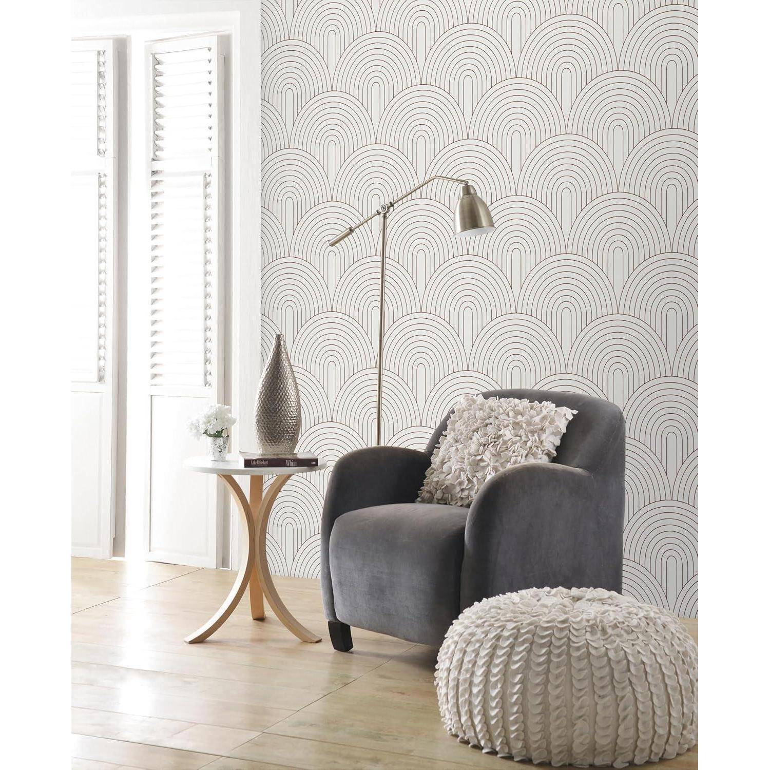Ein gemütliches Wohnzimmer zeigt einen grauen Sessel, einen runden Holzseitentisch mit einer Vase und einem Buch sowie einen weißen strukturierten Hocker. An der Wand befindet sich ein stilvoller, grauer Art-Déco-Muster.