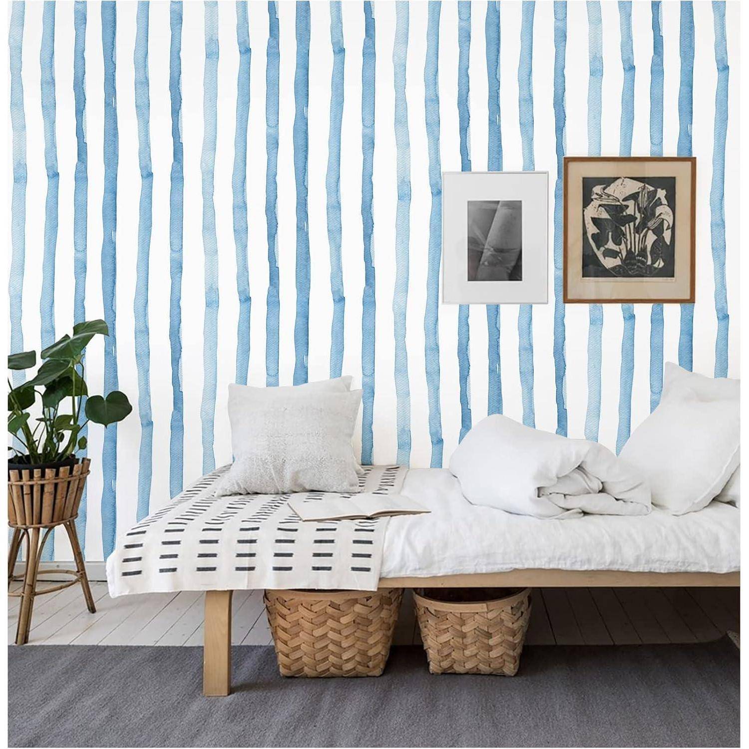 Ein gemütliches Zimmer mit einem weißen Bett, zwei Körben darunter und einer Pflanze links davon. Die Wand zeigt vertikale blaue und weiße Streifen und gerahmte Kunstwerke.